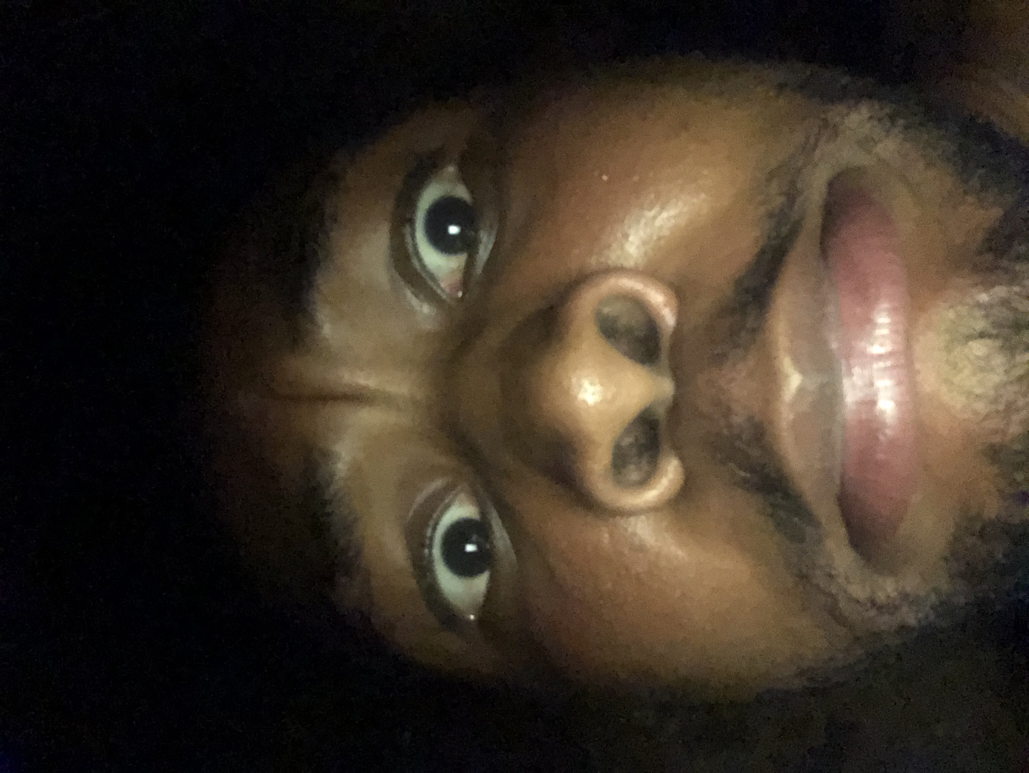 A close-up, low-angle photograph depicts a Black male's face. His eyes are open and looking upward, and his mouth is slightly open, revealing part of his tongue. The background is dark and indistinct.