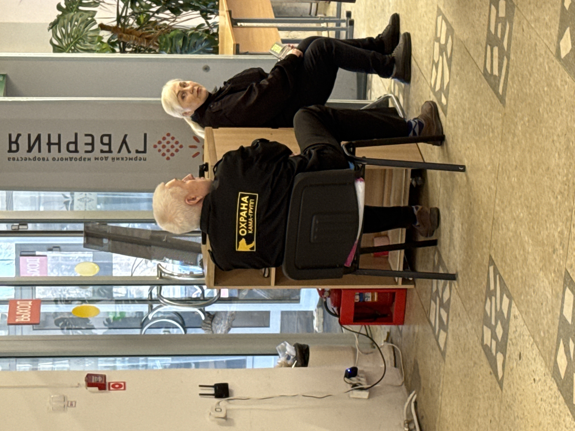 Two elderly individuals are seated in an indoor location in Perm, Russia. A fair-skinned woman with platinum blonde hair is sitting on a ledge, facing the camera. She is wearing black clothing and black shoes. Beside her, a fair-skinned man with white hair is seated in a black plastic chair, facing away from the camera. He is wearing a black jacket with yellow lettering in Russian that includes the word "OXPAHA" (security). He is wearing blue and purple argyle socks. In the background, there is a large sign in Russian. A window is visible to the left and a red fire extinguisher is next to the chairs. The floor is light-colored with a geometric pattern.
