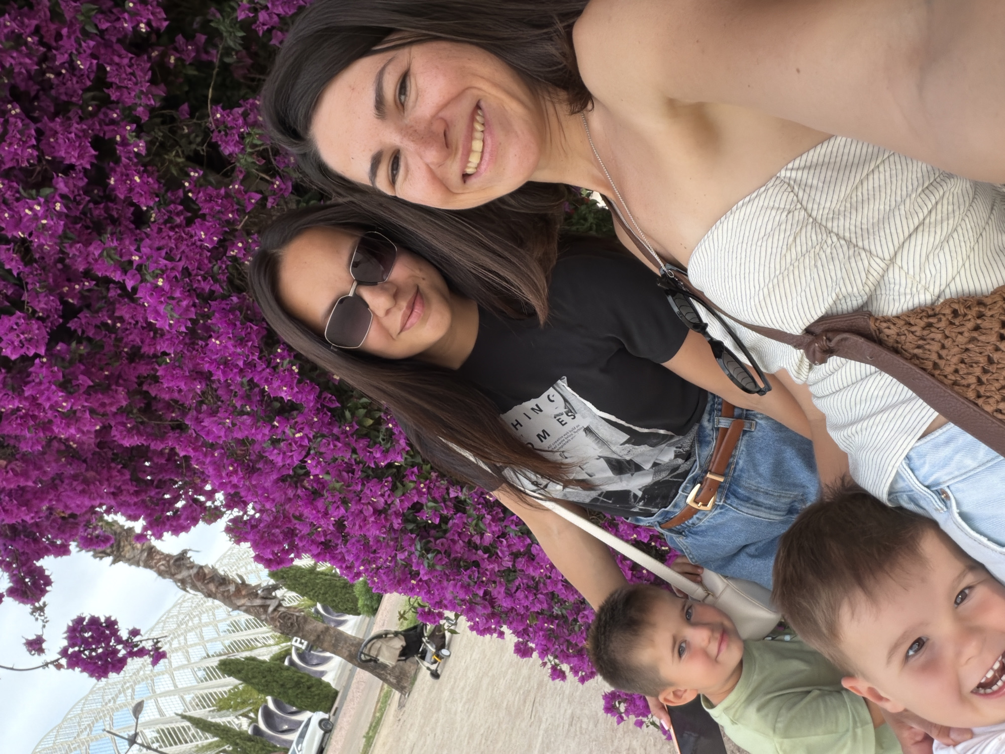 This is a selfie of two women and two young boys in front of a vibrant purple bougainvillea plant, with a modern white building visible in the background. The woman on the right is smiling broadly at the camera, wearing a strapless white and blue striped top and a silver necklace. She has freckles on her face. The woman on the left is wearing sunglasses, a black t-shirt with a graphic print, and denim shorts with a brown belt. She is looking towards the camera with a slight smile.
Two young boys are positioned in the foreground. The boy on the left is wearing a light green t-shirt and looking towards the camera with a calm expression. The boy in the very front is leaning in, smiling widely with his mouth open, revealing his teeth. He appears to be resting his head on the shoulder of one of the women.
The background reveals the distinctive architecture of the City of Arts and Sciences in Valencia, Spain. The white, futuristic building with its skeletal design is a prominent landmark. The purple bougainvillea is lush and abundant, framing the subjects. A palm tree trunk is also visible, and to the left, a paved walkway with trees and a stroller can be seen. The scene suggests a pleasant outdoor setting in Valencia.
