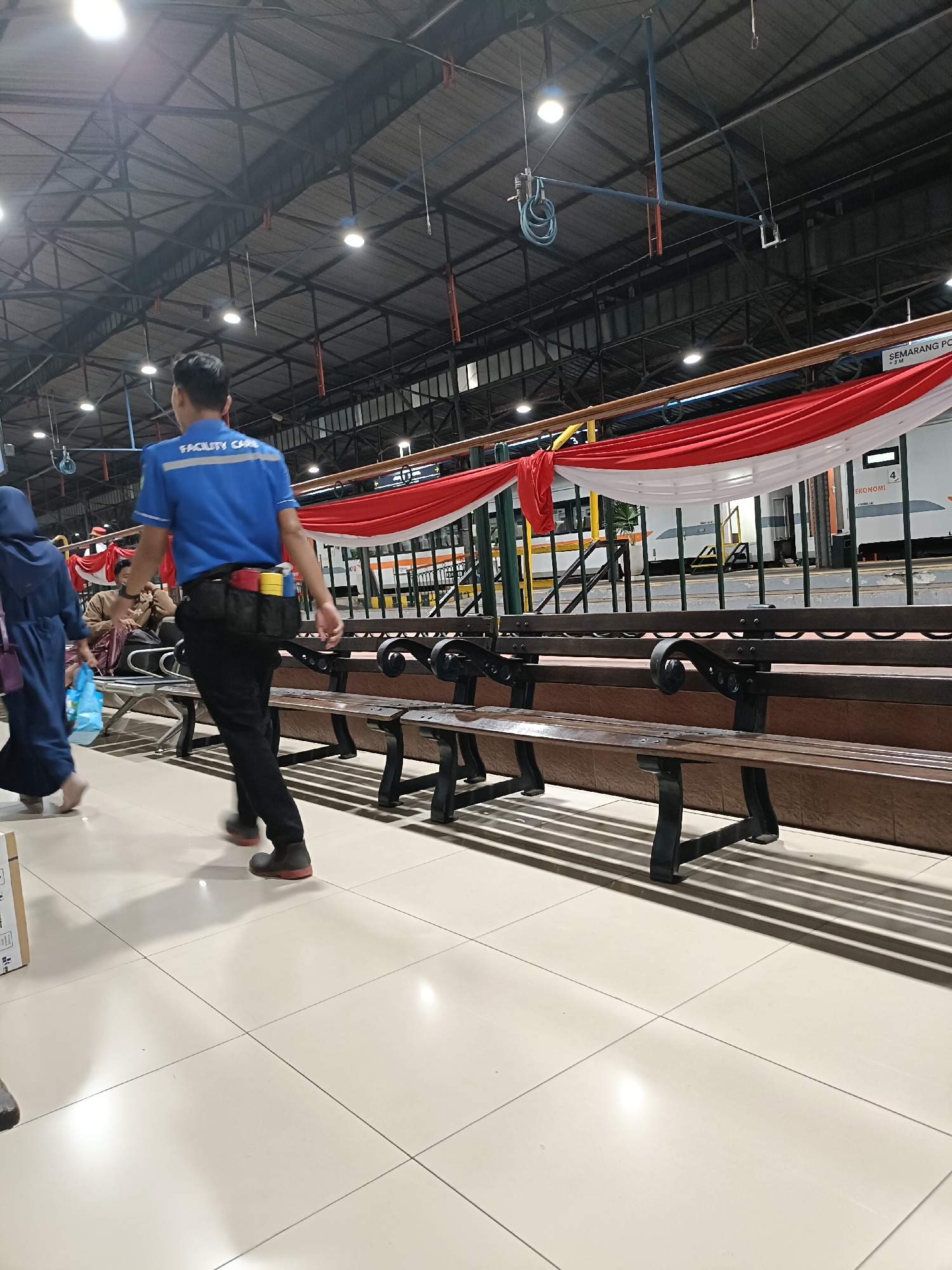 The image depicts the interior of a train station in Semarang, Indonesia, likely on a platform or waiting area. A person wearing a blue shirt with "FACILITY CARE" written on the back is walking to the right. They are wearing black pants and dark shoes with red soles. A utility belt is around their waist, containing various items. To the left, a person in a dark blue hijab and robe is walking away from the camera. In the background, there are rows of wooden benches with metal accents. A white and red Indonesian flag is draped along a railing above the benches. A train with the word "EKONOMI" visible on its side is parked at a platform behind the railing. The ceiling of the station is visible, supported by industrial beams and lights. The floor is tiled in a light color.
