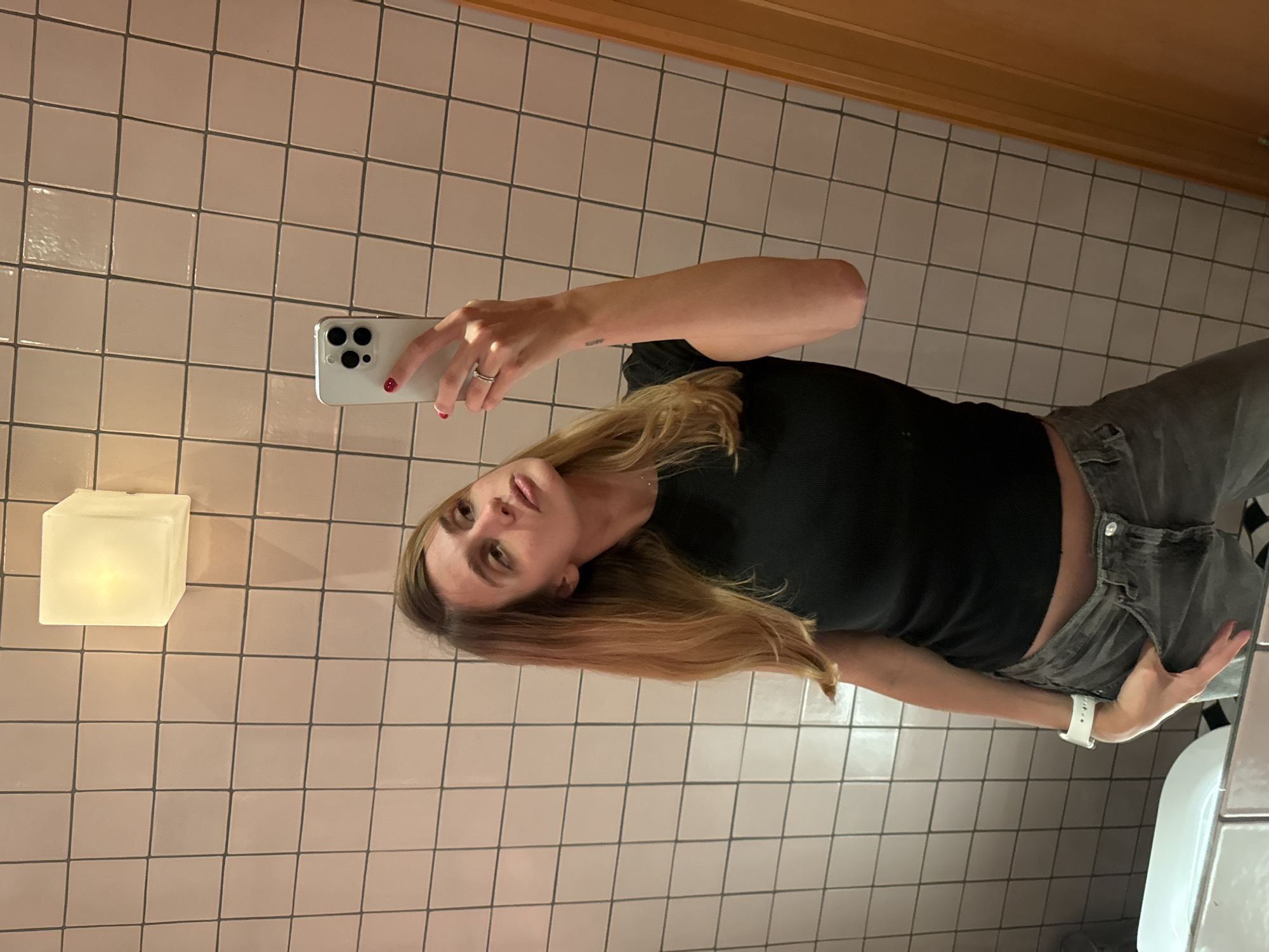 The image depicts a woman taking a selfie in a tiled bathroom. She is holding a smartphone in her right hand, her hair is shoulder-length and blonde, and she is wearing a black short-sleeved top and grey jeans. A white watch is visible on her left wrist. A square light fixture is mounted on the wall to the left. A toilet can be seen at the bottom right corner of the image. The bathroom walls are tiled in a grid pattern with pink tiles. No identifying information in the photograph indicates that the location is Yerevan, Armenia.