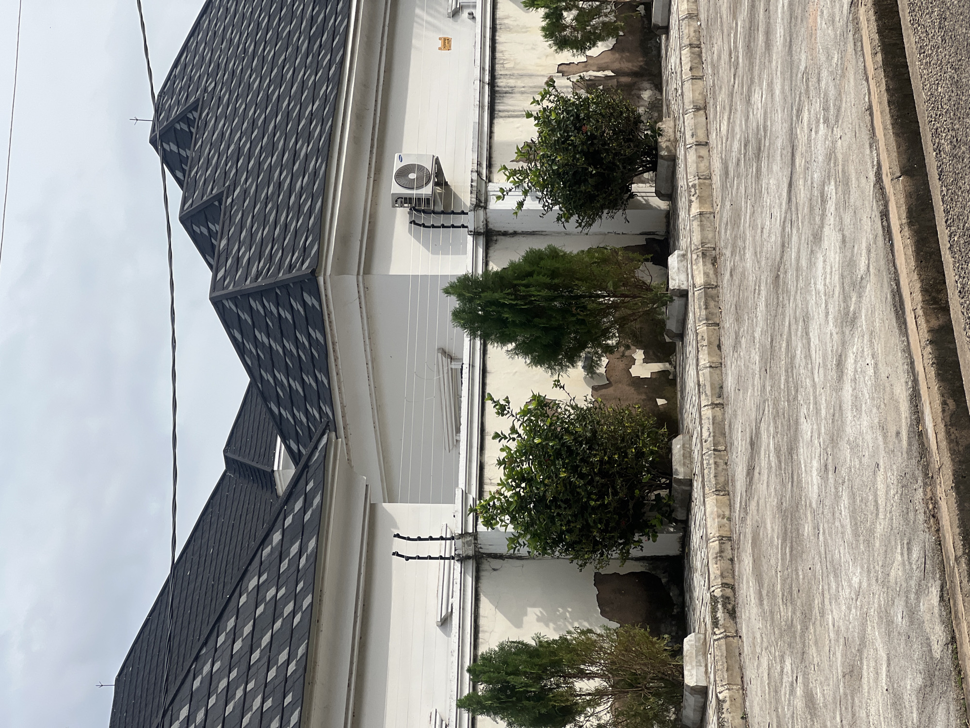 The image shows a section of a white building with a dark asphalt shingle roof, located in Jalingo, Nigeria. The building features an air conditioning unit mounted on the exterior wall. A security fence with multiple wires is installed along the top of the wall. In front of the building, there is a paved area with a curb, and several potted plants and trees are arranged along the wall. The ground in the foreground is also paved, with a drainage ditch along the edge. The sky is overcast.