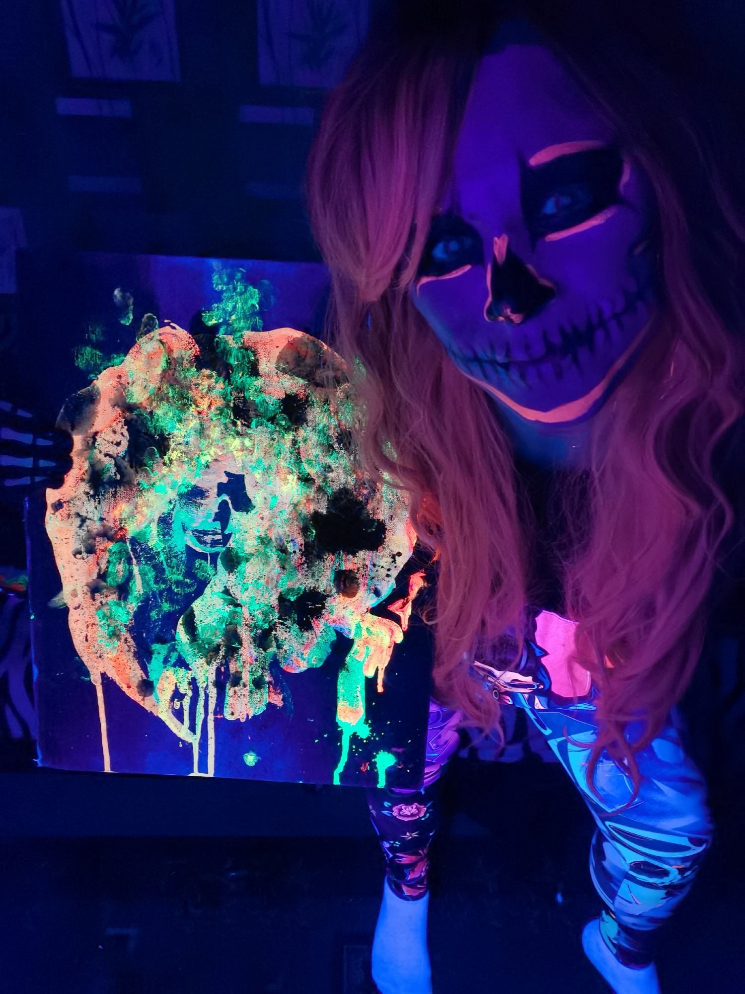 This image, taken in Chaikovsky, Russia, features a person in a dimly lit indoor environment illuminated by blacklight. The subject has intricate, UV-reactive skull makeup covering their face, with prominent purple, black, and orange hues, and striking blue eyes. Their long, wavy hair appears a vibrant pinkish-orange under the blacklight.
The person is holding a square canvas showcasing an abstract painting. The artwork is characterized by thick, textured applications and drips of neon green, bright orange/yellow, and black paint, all fluorescing intensely under the UV light. The background is dark, but faint outlines of what appear to be cannabis leaf designs are visible on a wall, suggesting a specific thematic decor. The person is also wearing patterned leggings that react to the UV light with swirling blue and purple designs, and bright white socks or footwear.
The scene conveys an artistic and vibrant atmosphere, indicative of a blacklight art show, themed party, or creative performance. The person's pose and the presentation of the glowing artwork suggest an engagement with or presentation of their art. No discernible text is visible.