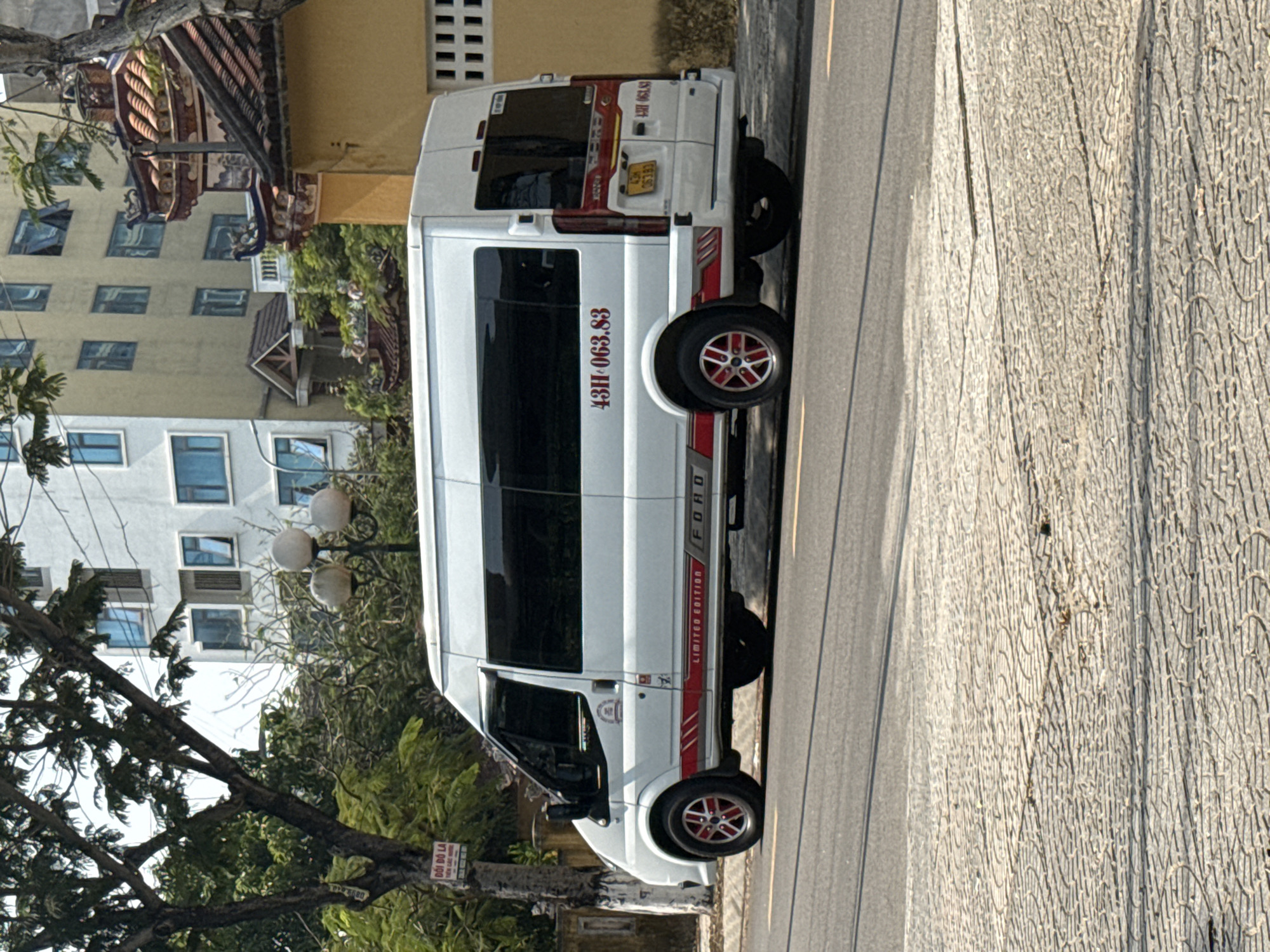 A white Ford Transit van with red accents and the license plate "43H-063.83" is parked on a street in Đà Nẵng, Vietnam. The van is a "Limited Edition" model. The background includes multi-story buildings with various architectural styles and a tree with a sign that reads "ĐỐI BÓ LA". The street is paved, and the lighting suggests it is daytime.