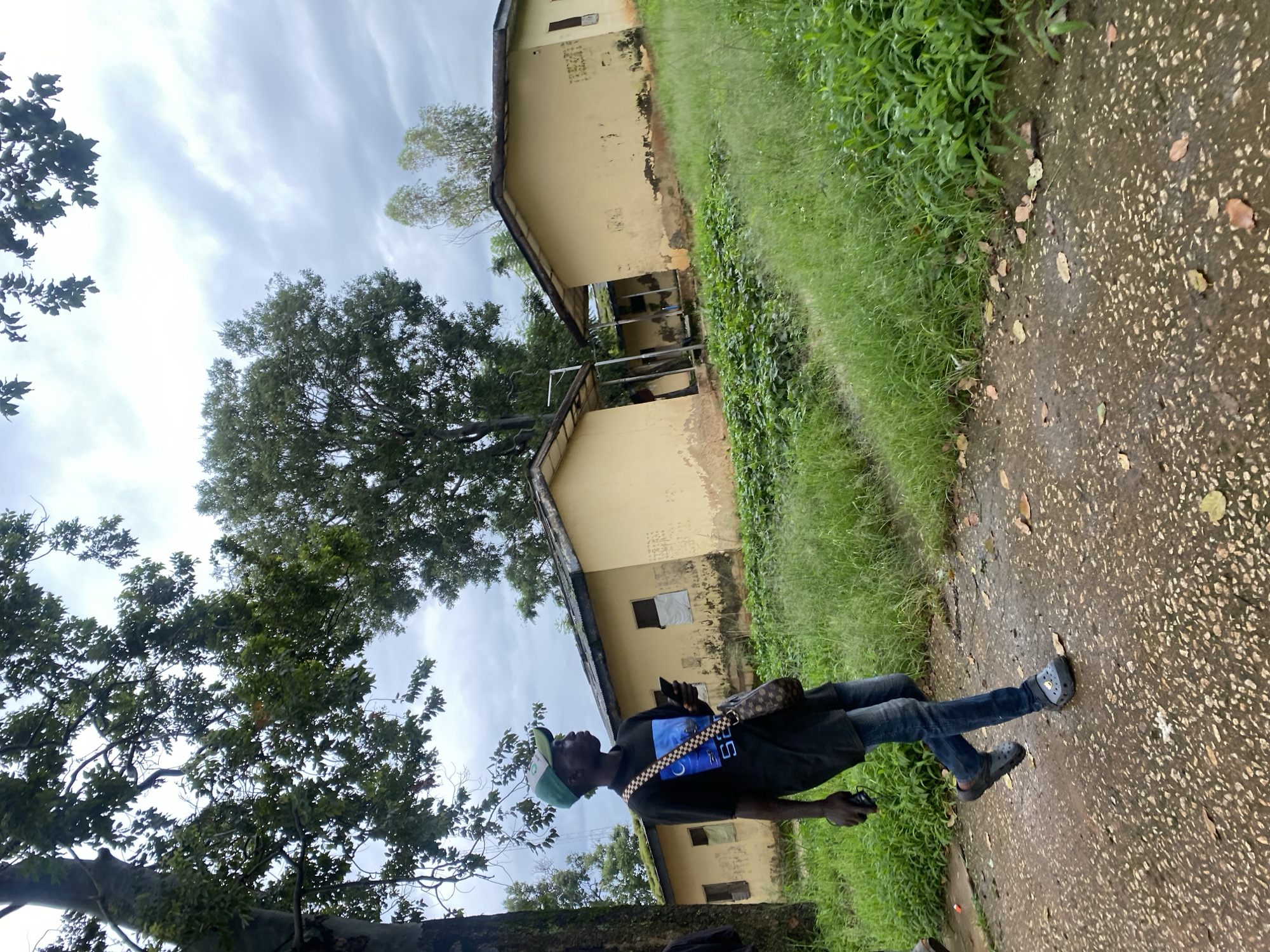The image shows a young man walking on a wet, pebbled path in front of two dilapidated, yellow buildings with corrugated iron roofs. The buildings appear to be small, single-story structures. Overgrown green grass and foliage surround the buildings and the path. Trees with dark bark and green leaves are visible in the background, with a cloudy sky overhead. The man is wearing a black t-shirt with a graphic design, blue jeans, a green baseball cap, and dark clogs. He is carrying a mobile phone in his right hand and a small bag with a patterned strap across his chest. The scene is likely located in a rural or semi-rural area within Jos, Nigeria, given the architectural style of the buildings and the vegetation.
