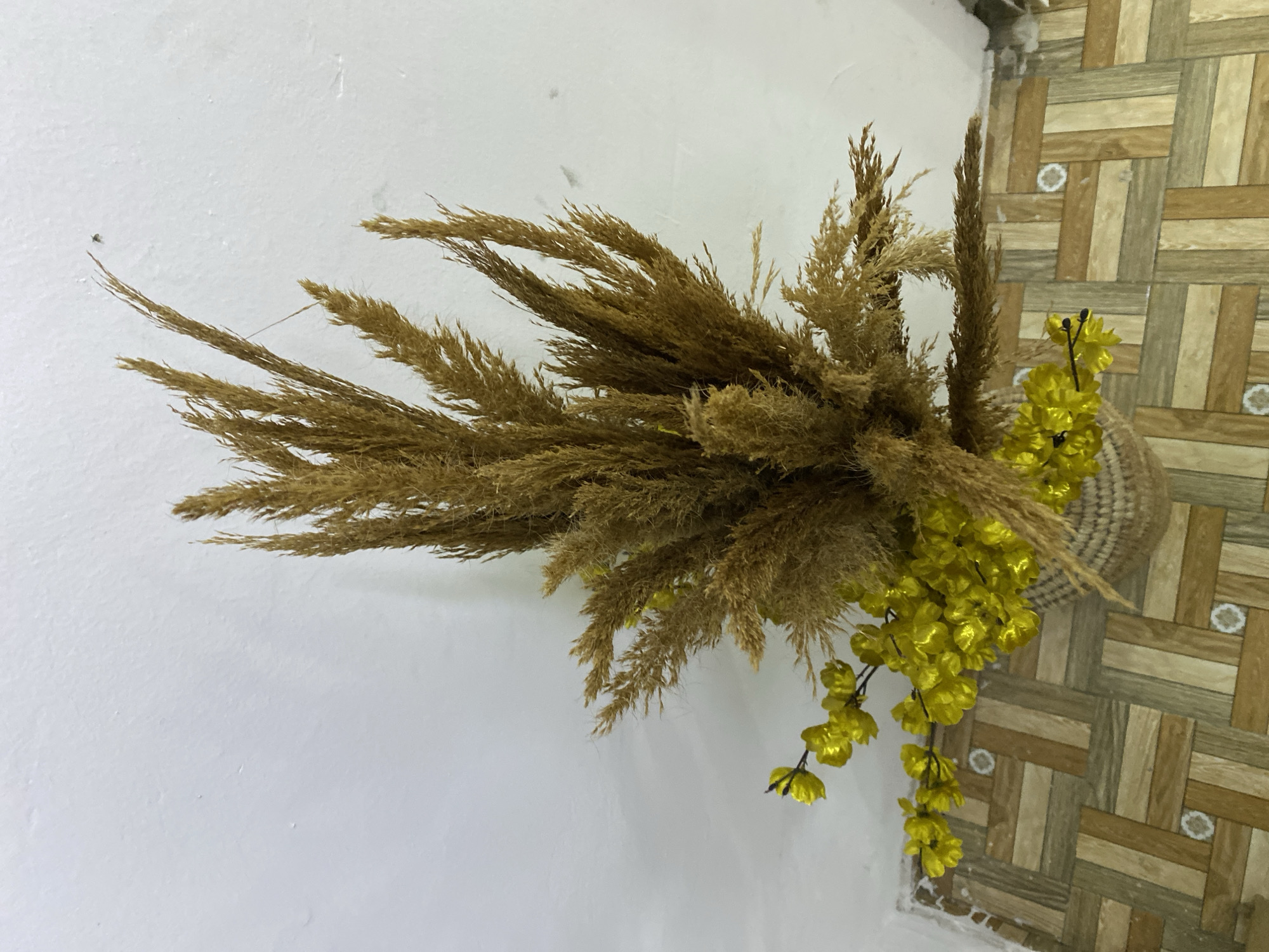 The image displays an indoor decorative arrangement featuring a prominent bundle of dried, feathery grass, likely pampas grass, in light brown and beige tones. Interspersed within the grass, particularly towards the right side, is a vibrant string of artificial yellow flowers. A woven basket or similar handcrafted item is partially visible behind the yellow flowers, suggesting it might be part of the display or serving as a container.
The background is divided into two distinct surfaces. On the left, a plain white wall provides a simple backdrop, showing some subtle texture and minor imperfections. On the right, a wall or floor exhibits a patterned design composed of varying shades of light and dark brown squares and rectangles, arranged in a basket-weave or parquet-like fashion. Some of the darker patterned sections include a subtle white circular motif.
There are no people, activities, or events depicted in the static image. The setting is an interior space, possibly a home or business in Badagry, Nigeria. The lighting suggests an indoor environment, but the specific time of day is not discernible. No visible text is present in the image.
