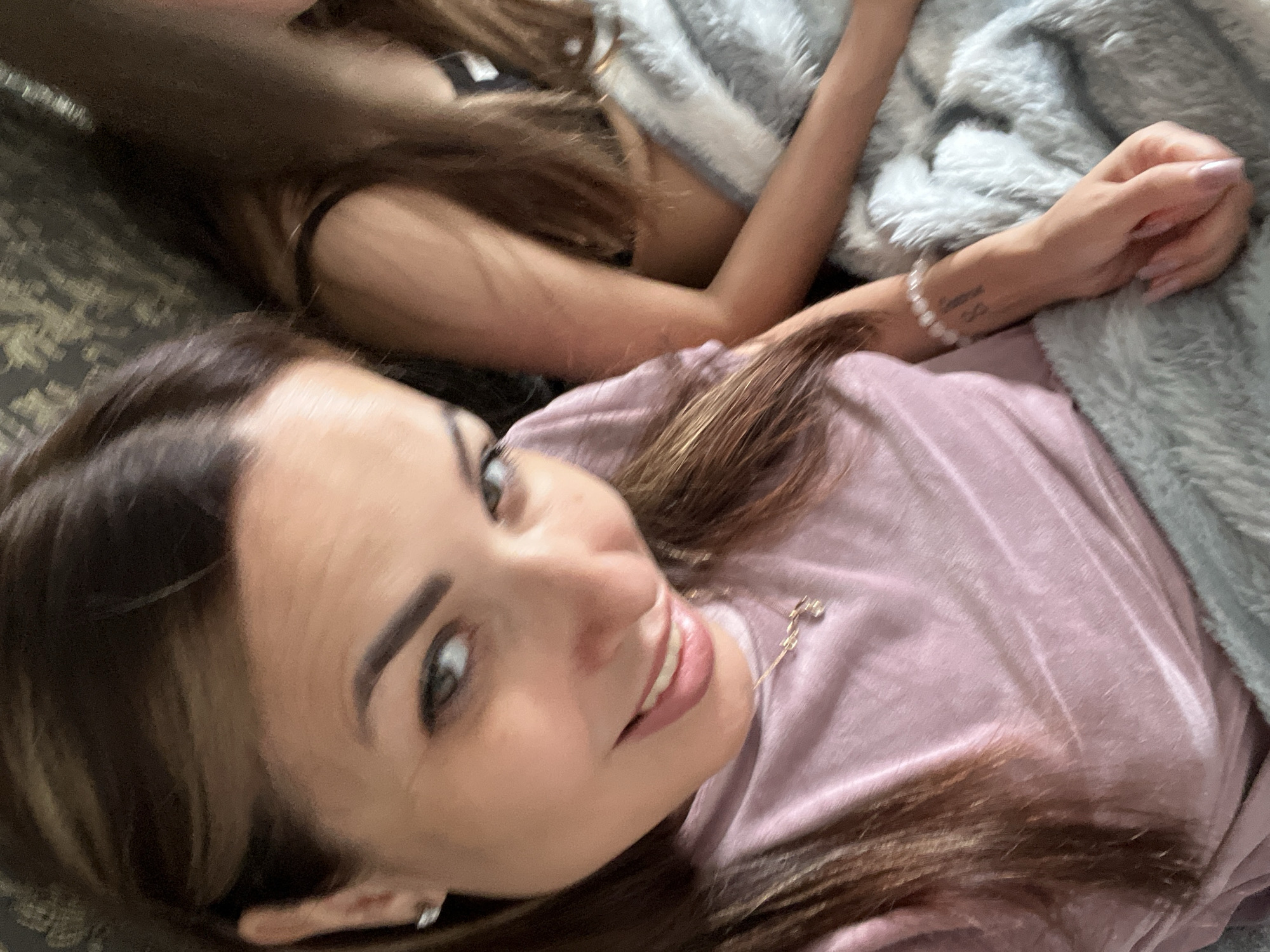 In an overhead shot, two individuals are positioned close together, partially covered by a soft, gray, fuzzy blanket. The person in the foreground, on the left, is a woman with brown hair, light skin, and wearing a pink, v-neck t-shirt. She is looking towards the camera with a smile, her head tilted slightly. Her right ear, adorned with a small, diamond stud earring, is visible. She is wearing a delicate necklace with a small charm. Her right arm rests on the blanket, and her hand is visible, with long, light pink fingernails. A bracelet of small, white beads is on her wrist, and a tattoo is visible on her forearm.
To her right and slightly behind her, another individual is partially visible. This person has long brown hair and is wearing a black top with thin straps. Their right arm is extended and also resting on the blanket. The background consists of a dark, textured surface, possibly a couch or bedspread. The lighting is soft, suggesting an indoor setting.