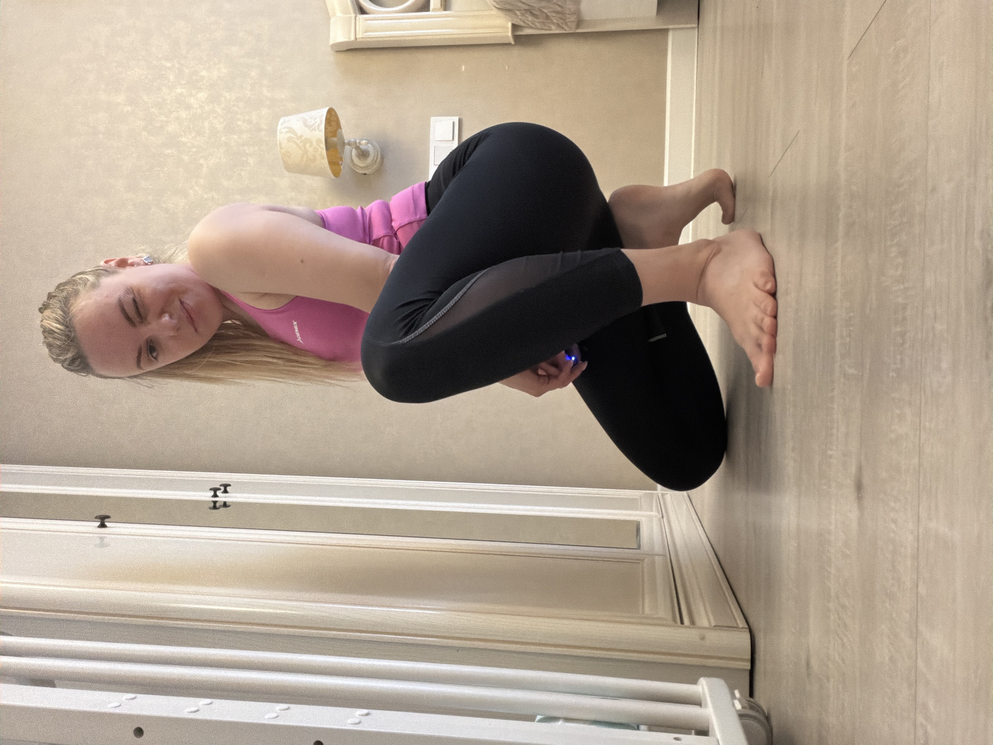 An indoor scene, likely in Votkinsk, Russia, features a young woman with blonde hair striking a flexible, seated pose on a light-colored wooden floor. Her hair is styled with prominent braids near the scalp, with the rest flowing down. She is dressed in athletic wear, consisting of a bright pink sports top, which bears the text "Xarex" (or similar brand name) on the chest, and black leggings with translucent mesh panels on the thighs. The woman looks directly at the camera, winking playfully, conveying a cheerful and confident demeanor. Her legs are intricately crossed and tucked, and she appears to be holding a small object that emits a soft blue glow between her hands.
The background consists of a light-toned wall with a subtle textured or patterned finish. A classic-style wall sconce with a decorative, patterned shade is mounted on the wall, alongside a white light switch or power outlet. White decorative moldings are visible towards the bottom of the wall, enhancing the room's aesthetic. In the lower left foreground, a section of white, slatted furniture, possibly a crib or bed frame, is partially visible. The overall lighting is soft and even, suggesting a comfortable and well-lit interior space.