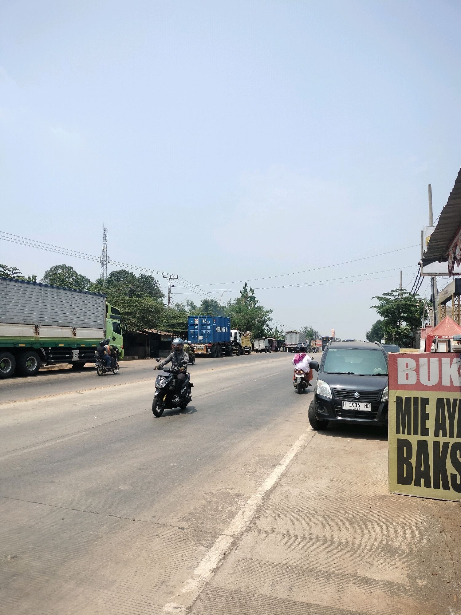 A bright, clear day is depicted on a two-lane asphalt road in Kutosari, Indonesia. The road runs horizontally across the frame, with a concrete shoulder on the right side. In the foreground, a black Suzuki Wagon R, license plate H 1016 HD, is parked partially on the shoulder. To its right, a large, vertical sign with yellow and black lettering advertises "BUKO MIE AYAM BAKSO."
On the road, a person wearing a helmet and a gray long-sleeved shirt rides a black motorcycle in the right lane, moving away from the camera. Another motorcycle, ridden by two individuals, is in the left lane, also moving away from the camera. Further down the road, a large, green and white semi-trailer truck with a silver cargo container is traveling in the left lane. Behind it, a blue shipping container truck is also visible.
The background consists of a line of trees and some low buildings. Overhead, power lines are strung across the sky. A tall telecommunications tower stands prominently in the distance, behind the trees. The sky is clear and light blue.