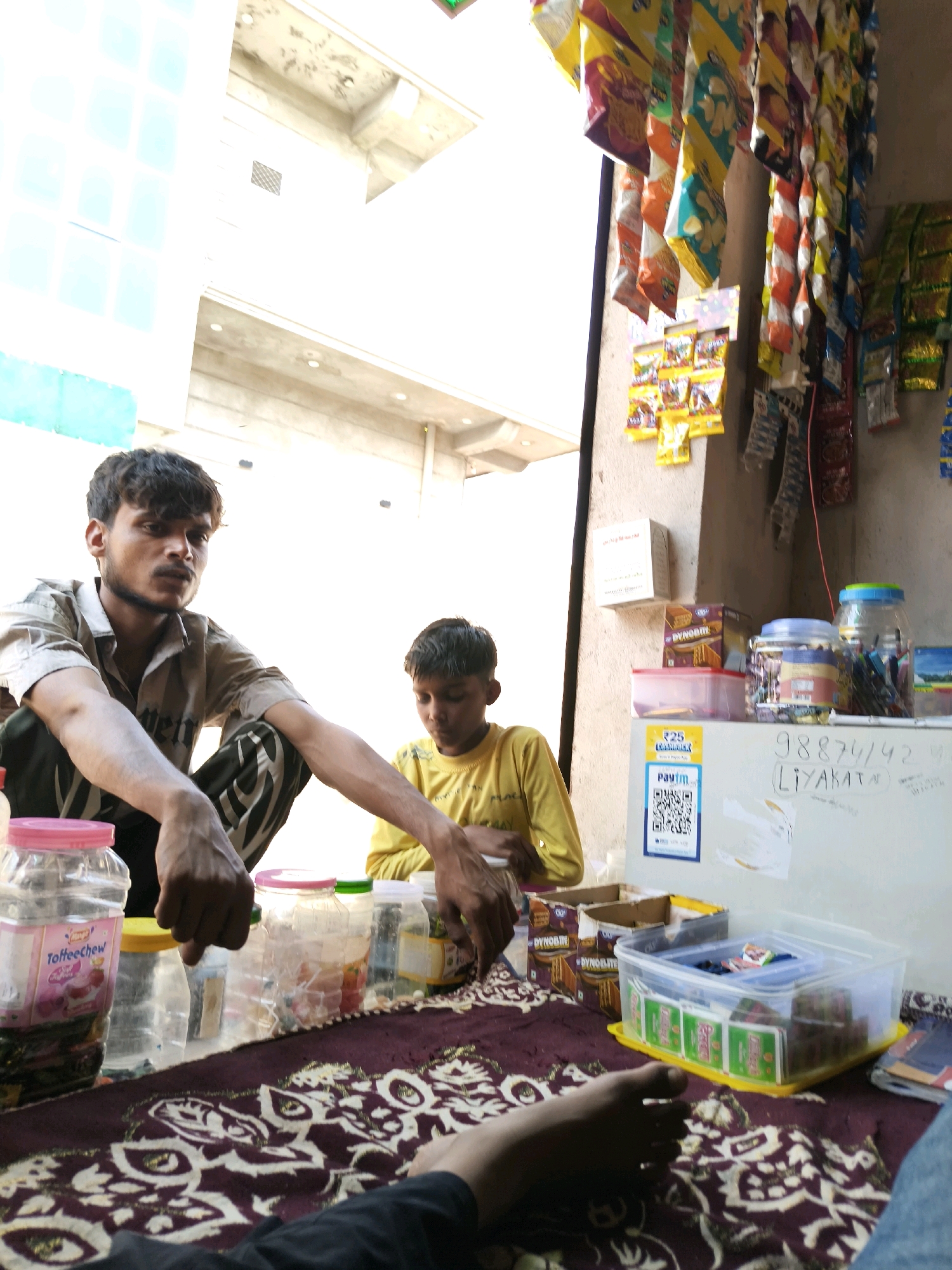 The image depicts an outdoor scene, likely a small shop or roadside stall, in Taranagar, India. Two individuals are present. A man in a light-colored shirt and dark pants is seated, looking towards the camera. Next to him, a younger person wearing a yellow shirt is also seated. The foreground shows a table covered with a maroon cloth featuring an intricate white pattern. On the table are various items, including plastic jars containing what appear to be snacks or candies, as well as some boxes. In the background, a wall is visible, adorned with packets of snacks hanging from above. A sign on the wall includes a QR code and what seems to be a phone number and a name written in Hindi. The overall impression is that of a humble commercial establishment.