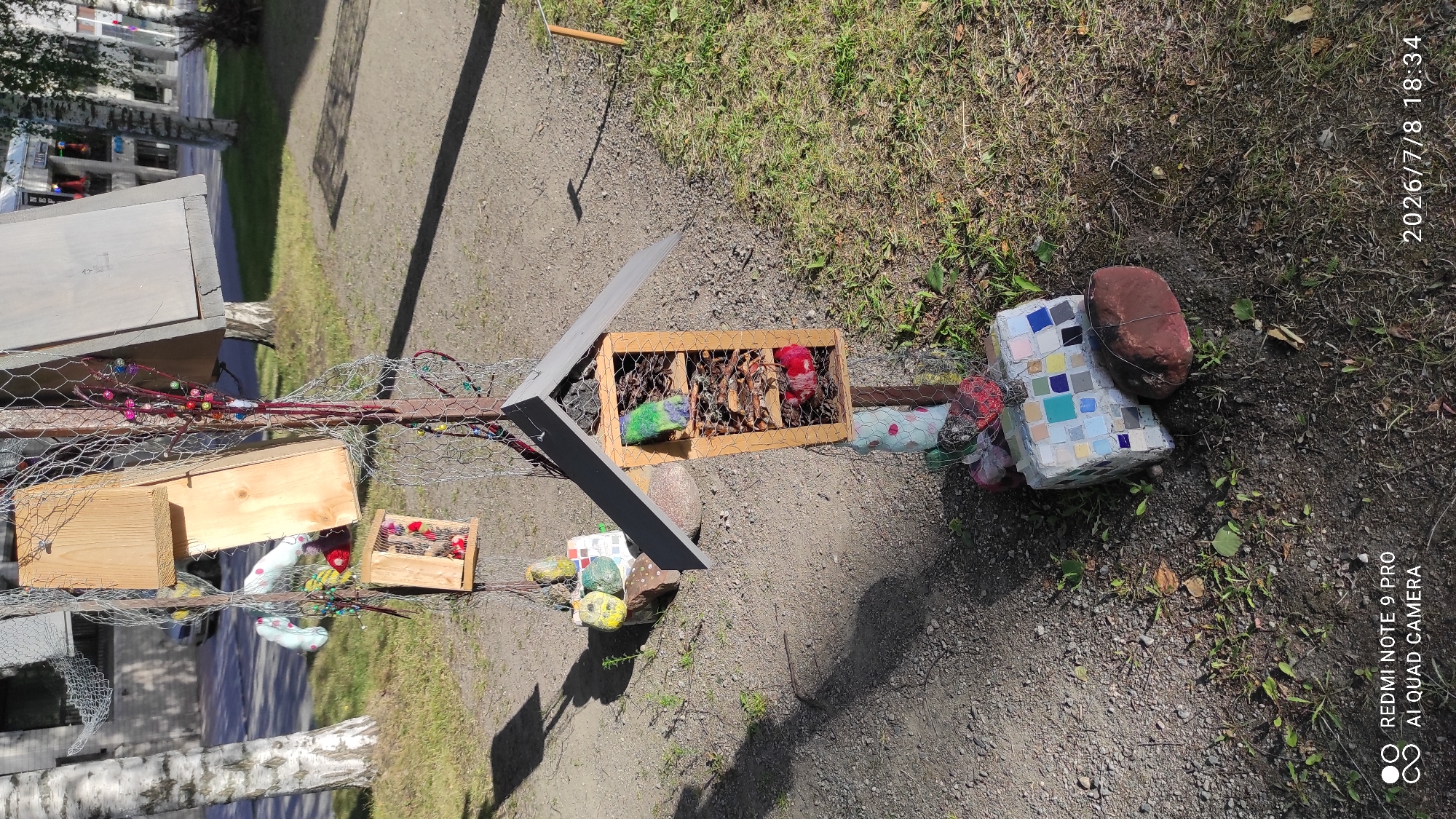 This is a photograph taken outdoors on a sunny day in Iisalmi, Finland. The subject is a homemade insect hotel constructed from wood and wire mesh, attached to a metal pole. The insect hotel has several compartments filled with natural materials like twigs and leaves, and some compartments contain colorful stuffed objects. At the base of the insect hotel, there is a mosaic-covered block and a large, reddish-brown rock. The background shows a paved pathway, grass, trees, and a building. The photograph has a timestamp in the bottom right corner indicating "2026/7/8 18:34" and a watermark in the bottom left corner reading "REDMI NOTE 9 PRO AI QUAD CAMERA".