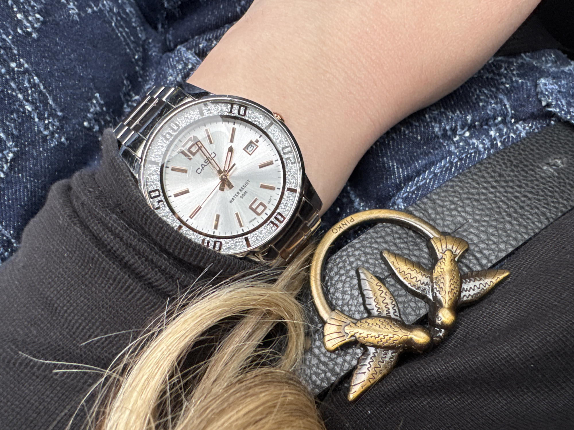 A close-up shot shows a woman's wrist adorned with a silver Casio wristwatch featuring a crystal-studded bezel. The watch face is silver with rose gold accents. A dark gray belt with a gold-colored bird-shaped buckle is positioned adjacent to the watch. Strands of blonde hair partially obscure the belt and watch. The woman is wearing denim jeans in the background. The scene does not contain sufficient information to determine its location in the city of Goritsa, Bulgaria.