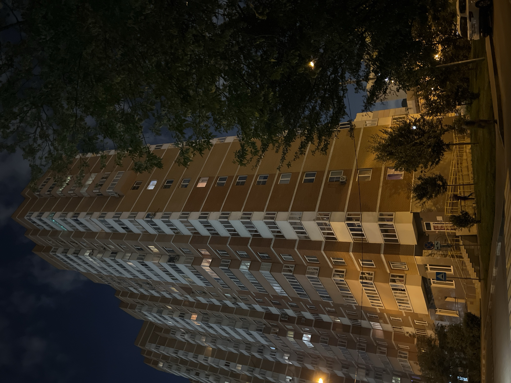 The image depicts a night-time view of a tall, multi-story residential building, likely an apartment complex, constructed with reddish-brown brick and white trim. The building features numerous windows and balconies, with some lights illuminated from within, suggesting occupancy. In the foreground, a street with a crosswalk is visible, along with a sidewalk and some greenery, including a large tree partially obscuring the building on the right. The sky is dark, indicating it is nighttime, with some clouds present. The lighting appears to be artificial, coming from streetlights and the building's interior. The scene suggests an urban environment, possibly a residential district within Moscow, Russia, characterized by dense housing and nighttime activity.