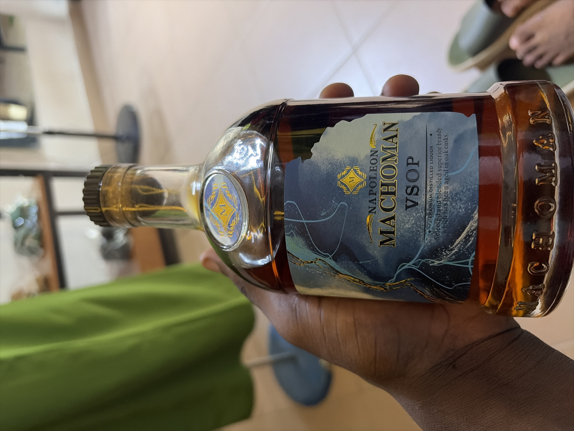 The image shows a hand holding a bottle of Napoleon Machoman VSOP brandy. The bottle is made of clear glass and contains a dark amber liquid. The label is predominantly dark blue with gold lettering. The text on the label reads "NAPOLEON MACHOMAN VSOP PREMIUM DISTILLED LIQUOR Selected and blended superior brandy which has been aged in oak casks." The bottle has a gold cap. The background is out of focus, but appears to be an indoor setting with a green surface and a light-colored wall. The lighting suggests it is daytime. The image is taken from a close-up perspective, focusing on the bottle and the hand holding it. The hand is holding the bottle at an angle, as if presenting it.