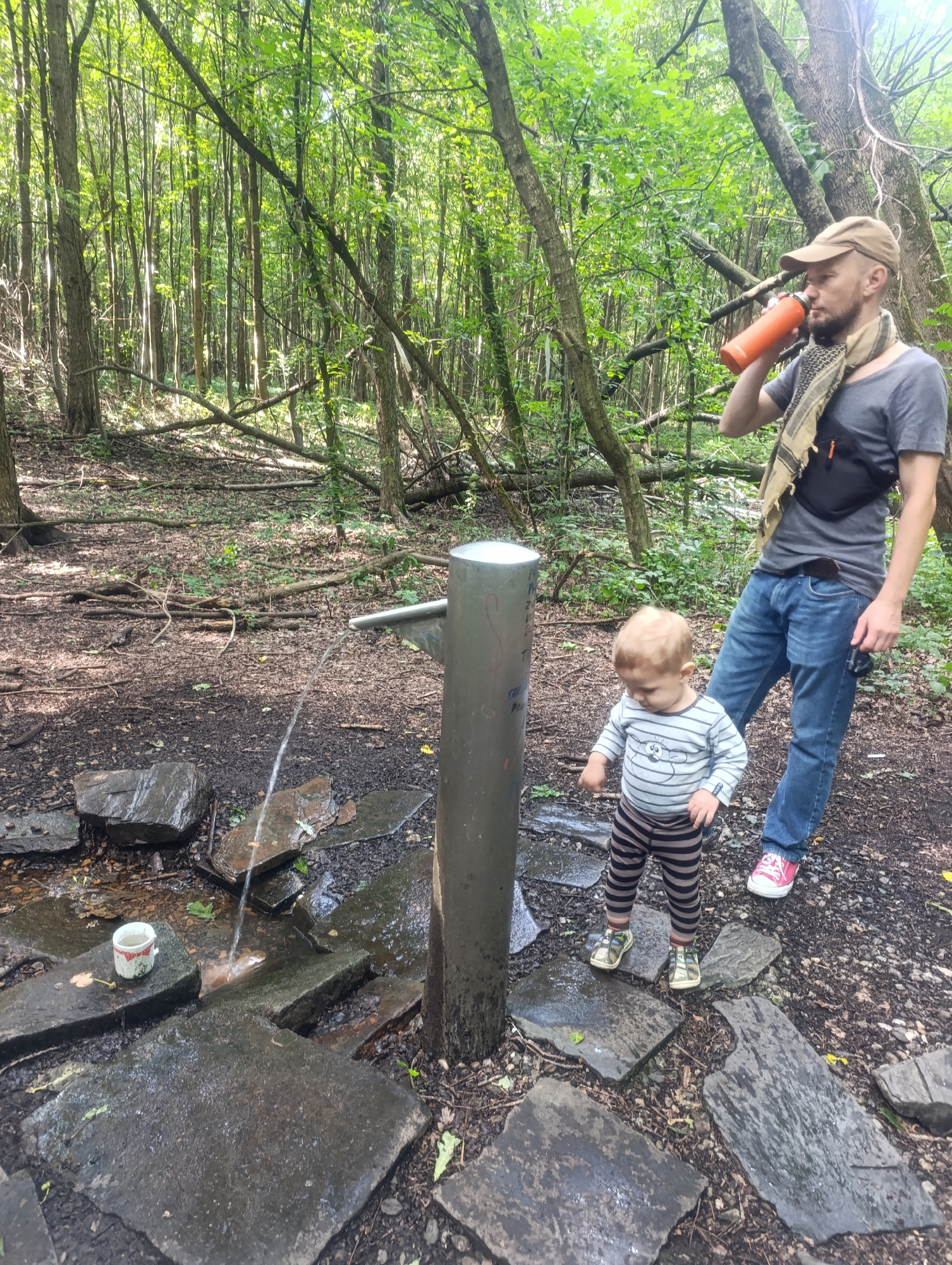 In a lush, sun-dappled forest setting in Olomouc, Czechia, a man and a young child are interacting with a natural spring. The man, wearing a tan baseball cap, a grey t-shirt, blue jeans, and a patterned scarf, is drinking from an orange water bottle. He has a black fanny pack around his waist. The child, dressed in a striped long-sleeved shirt and striped pants, stands near the spring, looking down at it with curiosity.
The spring is emerging from a metal pipe or faucet mounted on a cylindrical post. Water streams from the pipe, flowing over flat, dark stones that form a pathway. A small, white ceramic cup with red detailing rests on one of these stones near the spring. The forest floor is covered with fallen leaves, twigs, and tree roots, indicative of a natural, possibly untamed, environment. Tall trees with dense green foliage create a canopy overhead, allowing sunlight to filter through in patches. The overall mood appears peaceful and serene, suggesting a moment of shared exploration and refreshment in nature. The time of day appears to be daytime, likely late morning or afternoon, given the bright sunlight filtering through the trees. There are no visible indications of the weather, but the environment suggests it is likely warm and possibly humid. No discernible text is visible in the image.