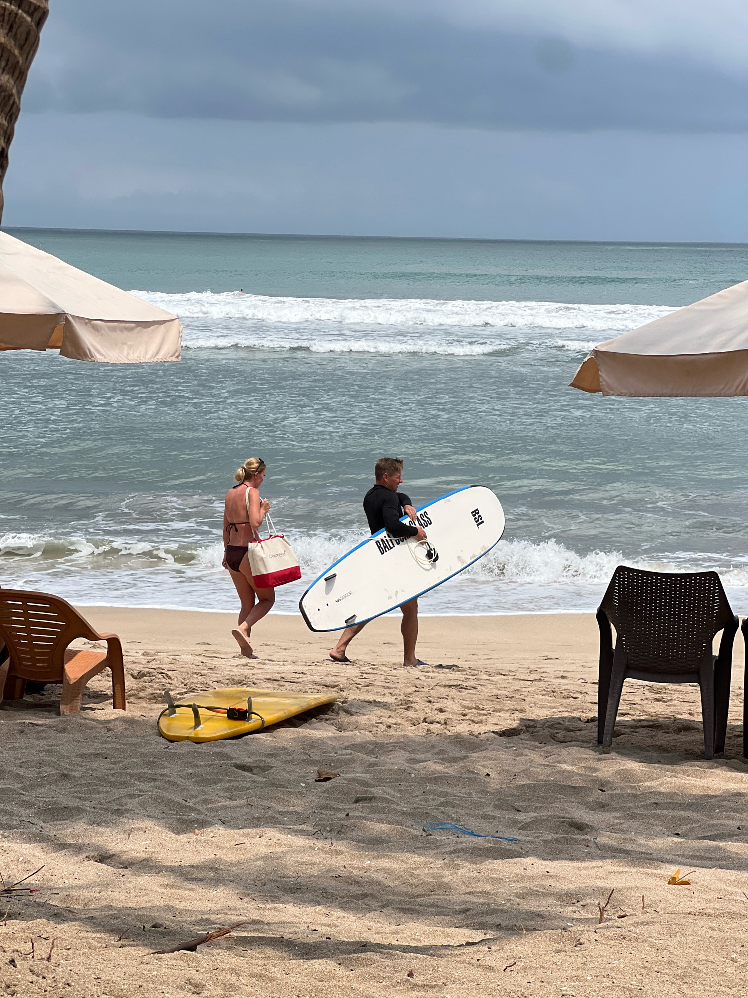 On a sandy beach in Kuta, Indonesia, two people walk towards the ocean. The person on the left, a woman, wears a dark bikini and carries a large tote bag with a red and white design. The person on the right, a man, is dressed in a black long-sleeved rash guard and shorts, and carries a white surfboard with blue trim. The ocean behind them has moderate waves with white foam. Two beach umbrellas are visible, one to the left and one to the right, casting shadows on the sand. A yellow surfboard lies on the sand in the foreground, to the left of the two walking individuals. Two chairs, one a light brown woven plastic and the other a dark woven plastic, are also present on the beach. The sky is overcast and gray.