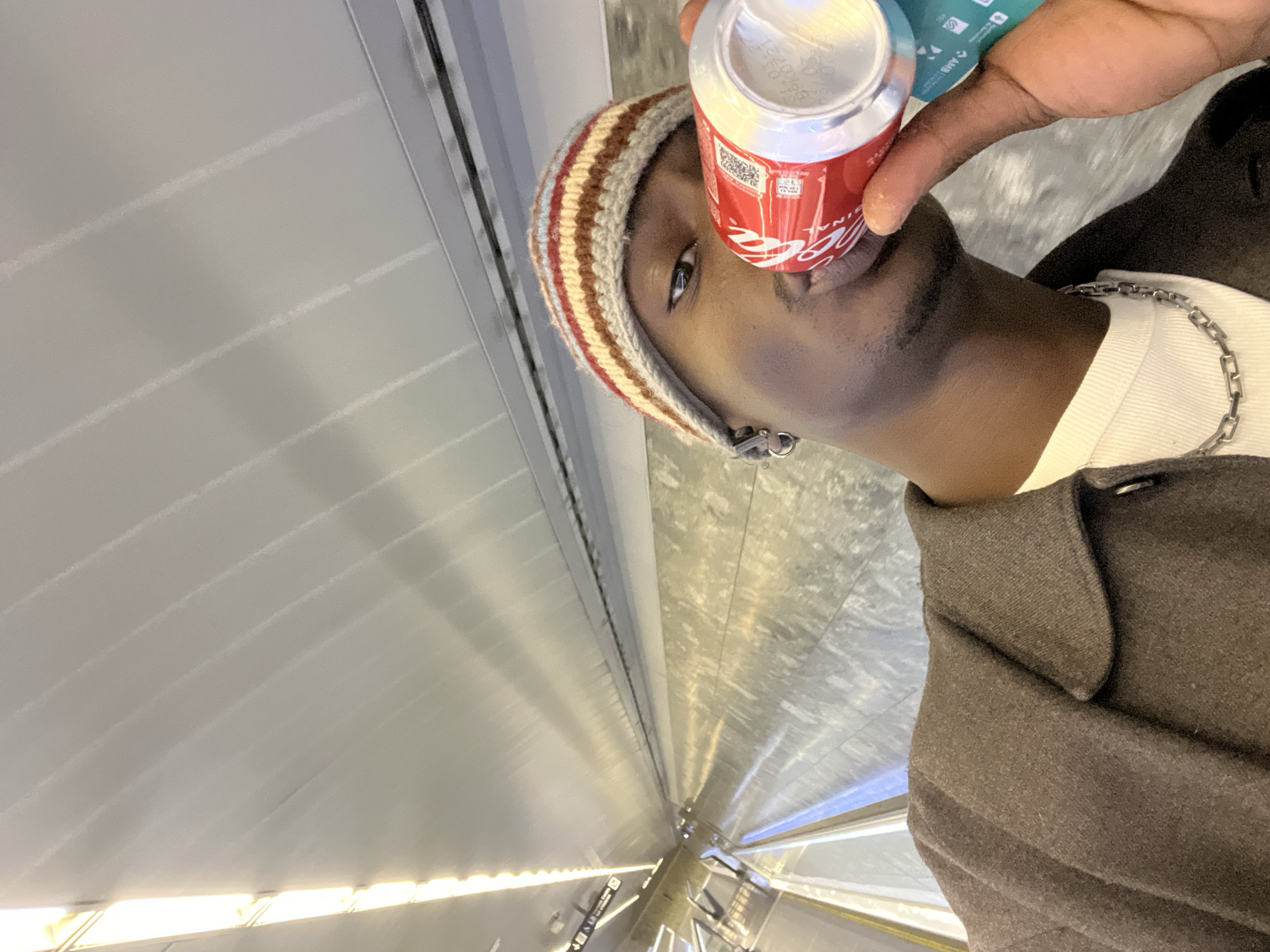 A person is drinking from a Coca-Cola can while standing in what appears to be a subway station in Barcelona, Spain. The person is wearing a brown and beige striped beanie, a white shirt, a silver chain, and a brown coat. They have an earring in their left ear. The subway station has white tiled walls and a bright light source from below.