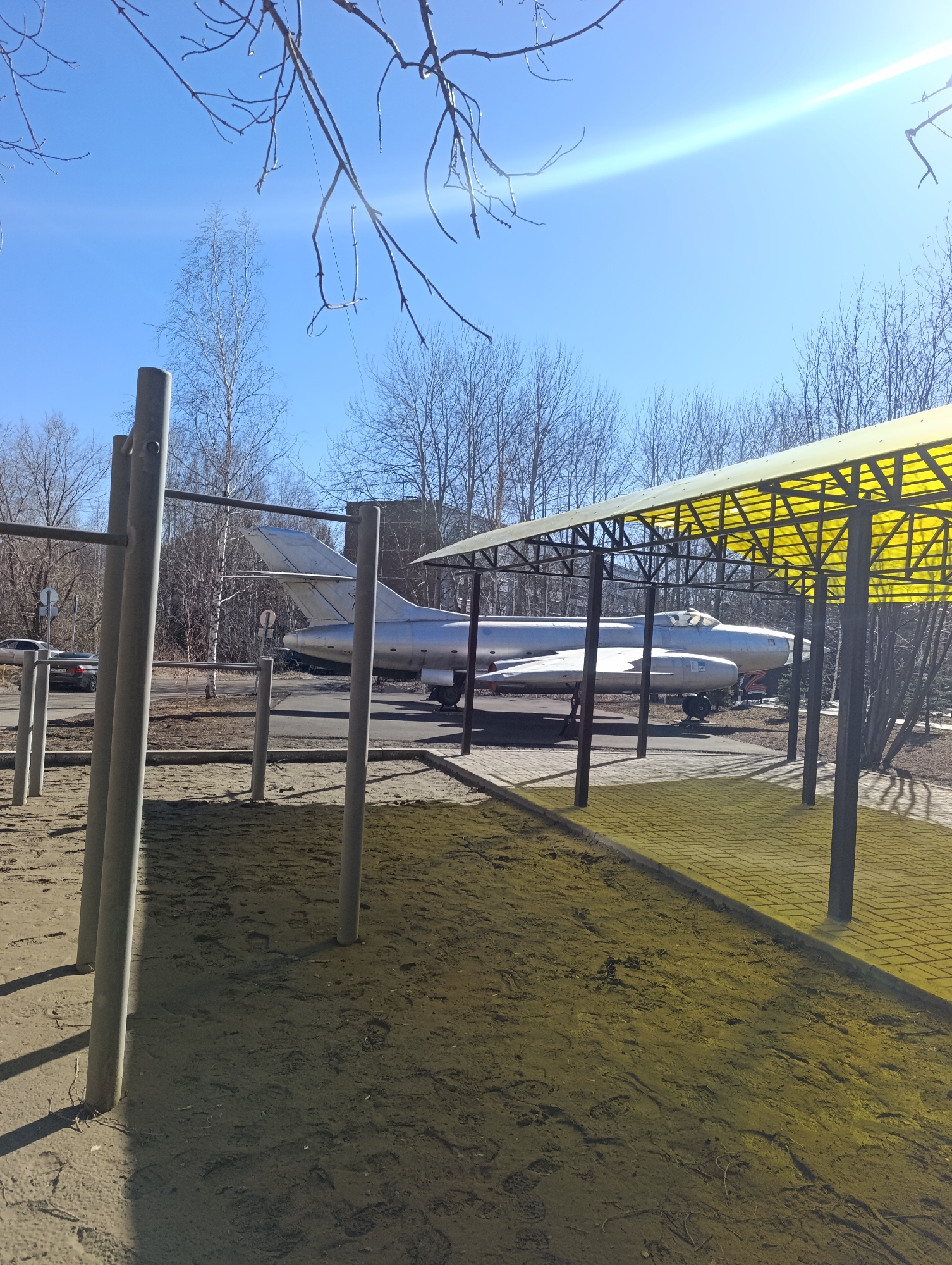 A silver Mikoyan-Gurevich MiG-21 fighter jet is displayed under a yellow-roofed shelter next to a street in Krasnoyarsk, Russia. The jet is surrounded by trees and is visible from behind a set of metal exercise bars in the foreground. The scene is illuminated by bright sunlight under a clear blue sky. A car is visible parked on the street.