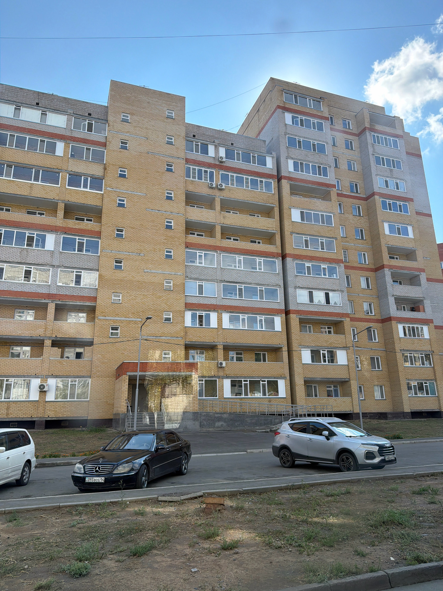 A long, multi-story apartment building made of yellow brick with grey accents and red horizontal bands is pictured under a bright blue sky with scattered white clouds. The building has numerous windows and balconies. In front of the building, a street is visible with two cars parked: a black Mercedes-Benz sedan and a silver SUV. The area in front of the building is a paved street with a sidewalk and a patch of dry grass and dirt. The image appears to be taken from a slightly elevated position. The license plates on both cars are visible and appear to be from Kazakhstan, with Cyrillic script. Based on the license plate format and the architectural style, the location is likely the city of Pavlodar, Kazakhstan.