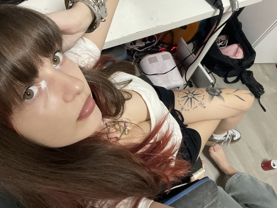 A woman with brown hair and striking green eyes gazes at the camera. She is wearing a white top with a glimpse of a necklace. A prominent tattoo is visible on her right thigh. The scene is cluttered with a desk, various wires, a bag, a foot in a shoe, and another barefoot foot. A can of soda lies on the floor. There is a distinct lack of organization.