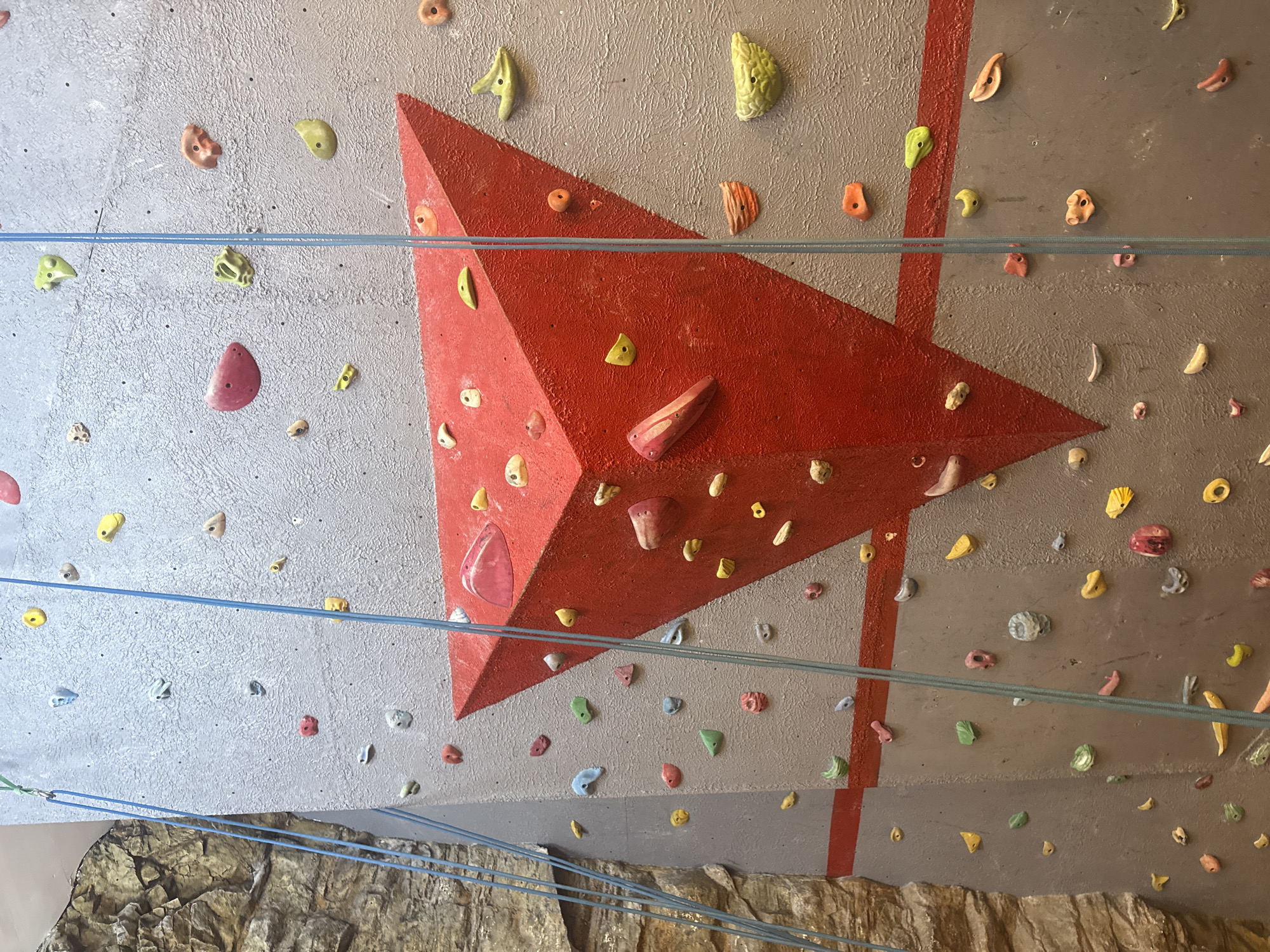 This media file presents an indoor climbing wall, likely situated in Puebla, Mexico. The wall features a textured, light grey background adorned with large, prominent red geometric volumes resembling pyramids or triangles, which add depth and varying angles to the climbing surface.
Numerous multi-colored climbing holds are bolted across both the grey and red sections of the wall. These holds vary greatly in shape, size, and color, including vibrant yellows, greens, pinks, oranges, blues, whites, and reds, indicating a diverse range of climbing challenges and routes.
Several blue ropes are strung horizontally across the wall at different heights, suggesting the setup for rock climbing activities such as top-roping or lead climbing. One rope at the bottom left is visibly attached to a metal anchor, extending towards a rugged, brown, rock-like formation that forms the base of the scene, possibly a padded landing area or part of the gym's architectural design.
The lighting is bright and even, consistent with an indoor climbing gym environment. No individuals are present in the image, but the arrangement of holds and ropes clearly indicates the space is prepared for climbing. There is no discernible text visible on the wall, holds, or ropes. The overall impression is that of a well-equipped facility ready for climbers.