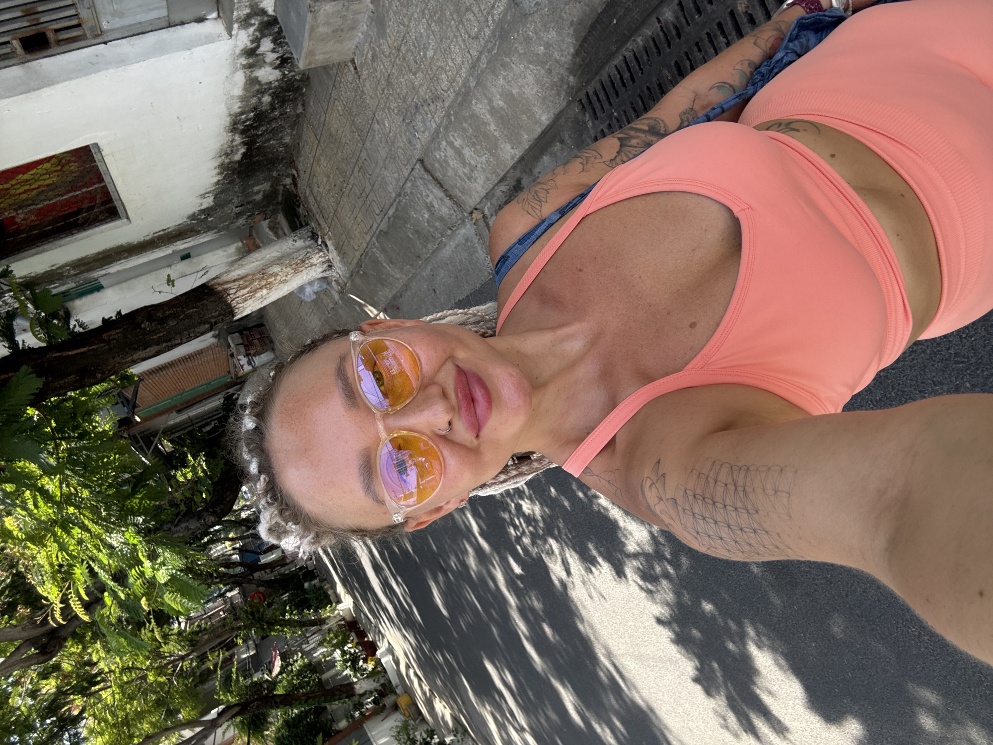 A woman with braided hair and a nose ring takes a selfie on a tree-lined street in Bac Nha Trang Ward, Vietnam. She is wearing a pink sports bra and leggings, and orange-tinted sunglasses. Her left arm is visible, displaying several tattoos. The street is paved, with a concrete sidewalk and a drainage grate on the right. Buildings with white walls and some signs of wear are visible on the right side of the street, while lush green trees line the left side. Shadows from the trees cast patterns on the asphalt.