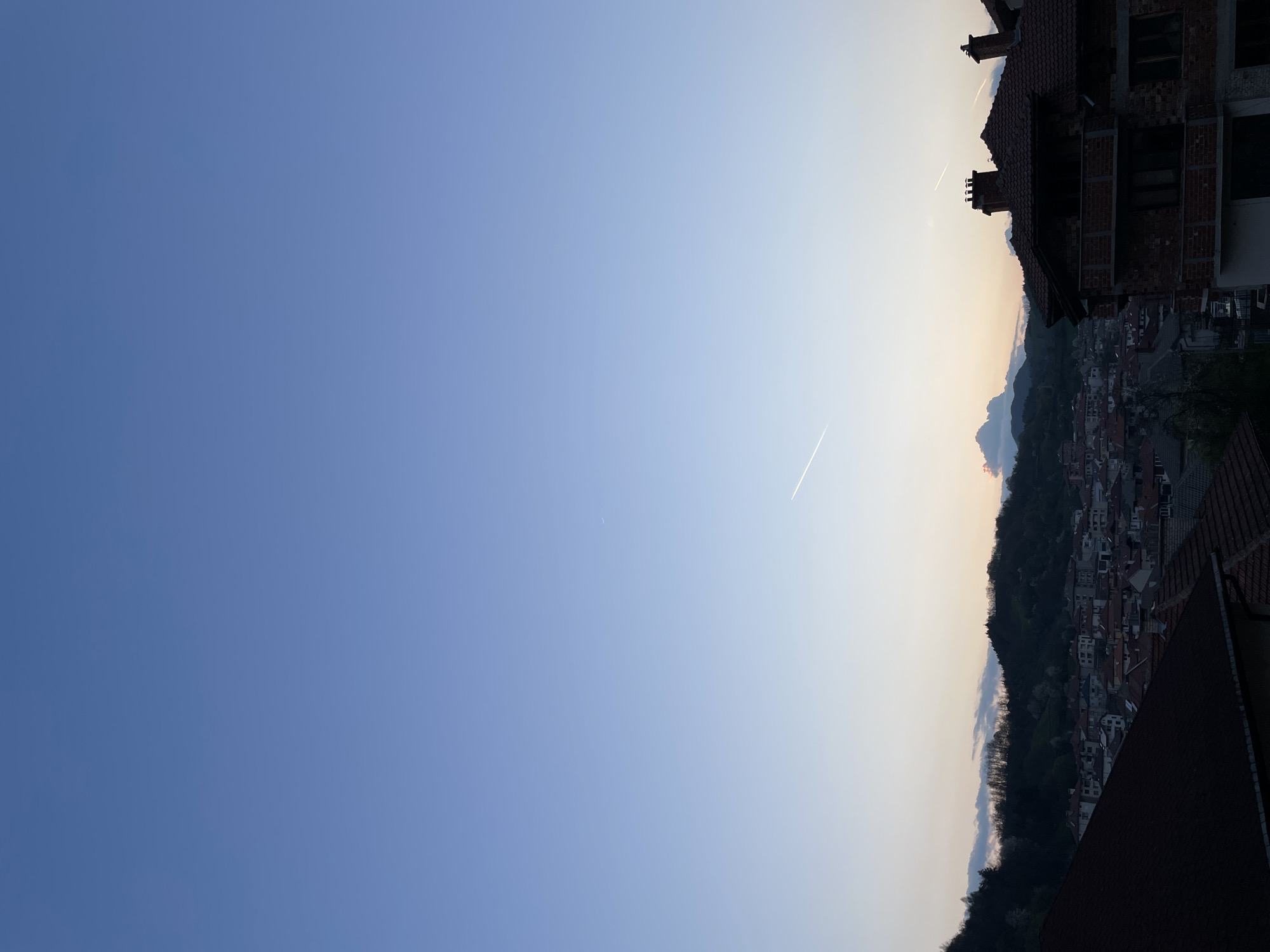 The image depicts a view of Zlatograd, Bulgaria at dusk. A clear blue sky transitions to a lighter hue near the horizon, with contrails visible from aircraft. The foreground contains buildings with red-tiled roofs and unfinished brickwork. Further into the distance, the town is nestled amidst green hills and mountains, with layers of buildings receding into the background. The light is soft and even, suggesting either dawn or dusk.