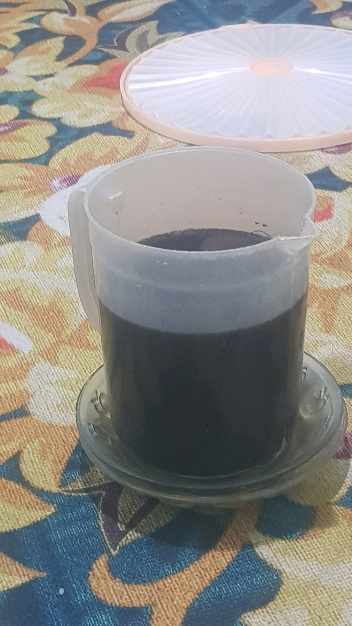 An overhead shot shows a plastic pitcher filled with a dark liquid, likely coffee, sitting on a clear glass saucer. The pitcher and saucer are placed on a patterned tablecloth with a floral design in shades of yellow, blue, and orange. In the background, a white plastic lid with a ribbed pattern is visible. The scene appears to be indoors, possibly in a home kitchen or dining area in Bakalan, Indonesia. There are no people or animals visible in the image.