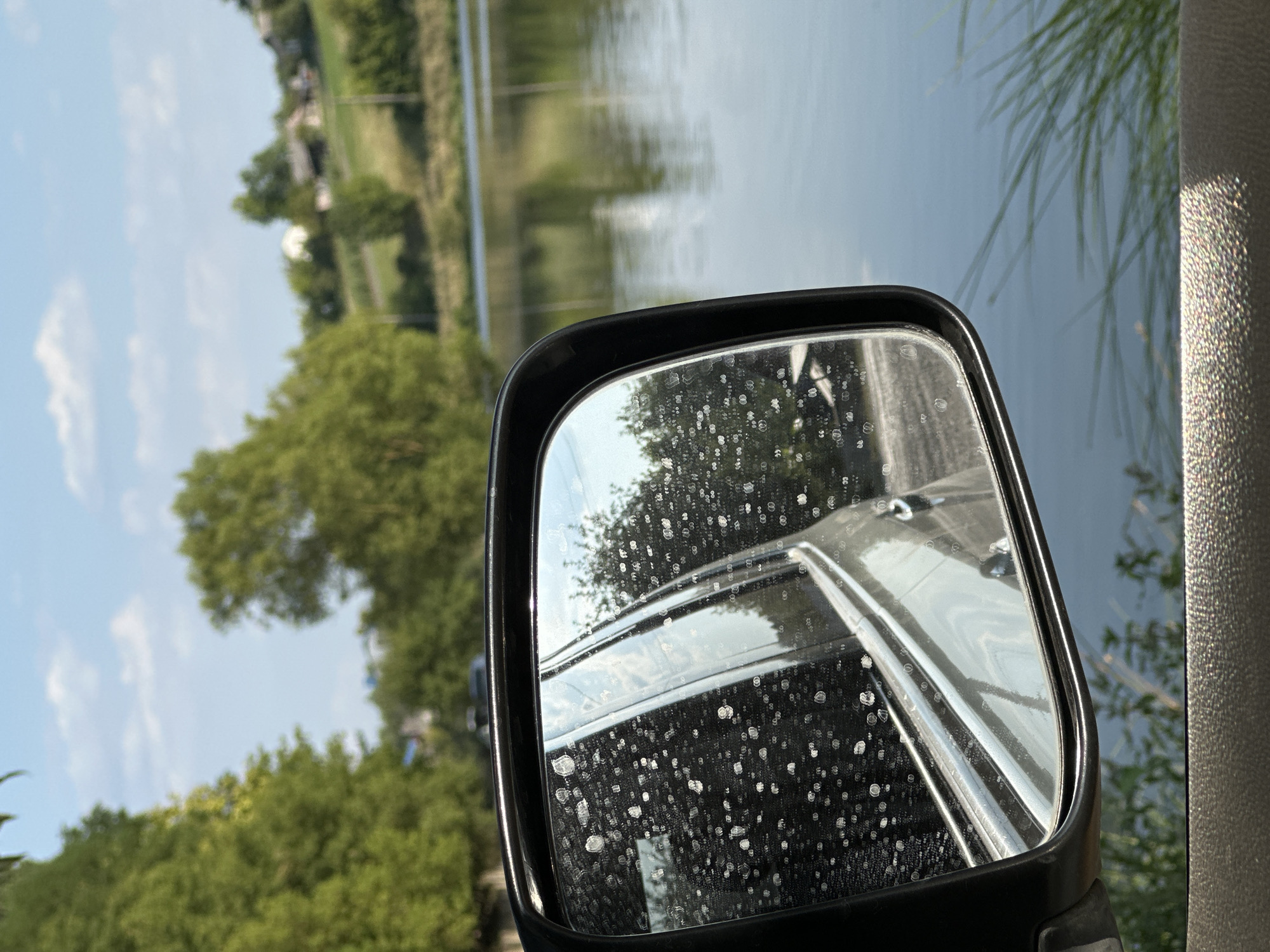 The image shows a side view mirror of a car parked near a body of water. The mirror reflects the car's interior and the surrounding landscape, which includes trees, houses on a hill, and the sky with clouds. The mirror itself is partially obscured by water droplets, giving it a speckled appearance. The water body appears to be a lake or a wide river, with a calm surface. In the foreground, there is a portion of the car's interior visible, with a textured dashboard. The overall scene suggests a rural or semi-rural setting. The prompt mentions the city of Makiv, Ukraine, but this specific location cannot be confirmed from the visual information alone.