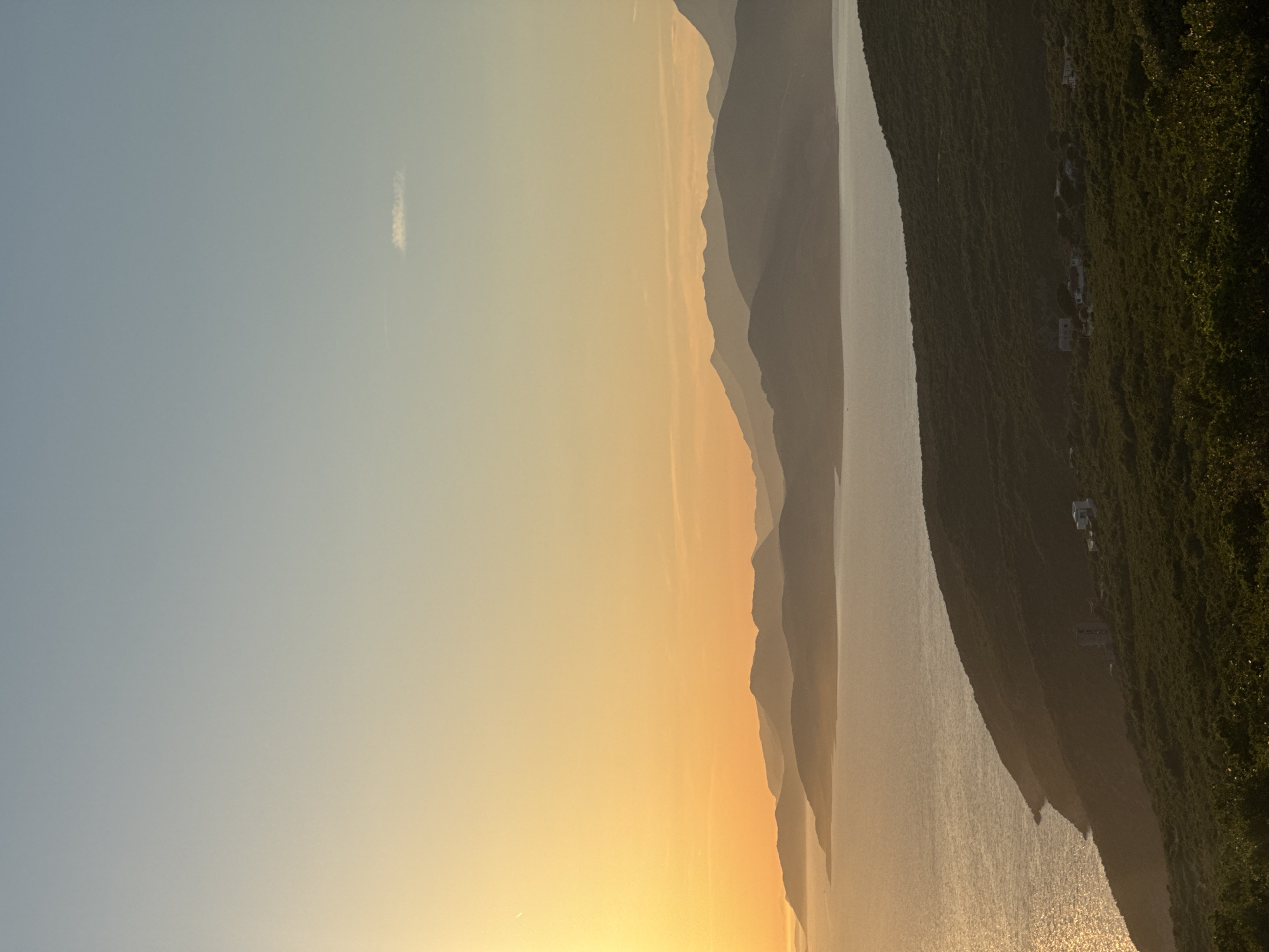 An aerial vertical shot captures a serene sunset over Kovačko Polje, Montenegro. The sun, a bright golden orb, sets ablaze the sky with hues of yellow and orange. A gentle sea laps at the shore, its surface shimmering with the sun's golden light. In the foreground, a lush green hill is dotted with a few sparsely placed houses, their architecture simple and functional. The background is dominated by a series of undulating mountains, their peaks fading into the hazy horizon, creating a sense of depth and grandeur. The sky transitions from a warm yellow at the horizon to a soft blue at the zenith, with a single wisp of cloud drifting lazily across the expanse. The scene evokes a sense of peace and tranquility, a perfect encapsulation of a Mediterranean evening.