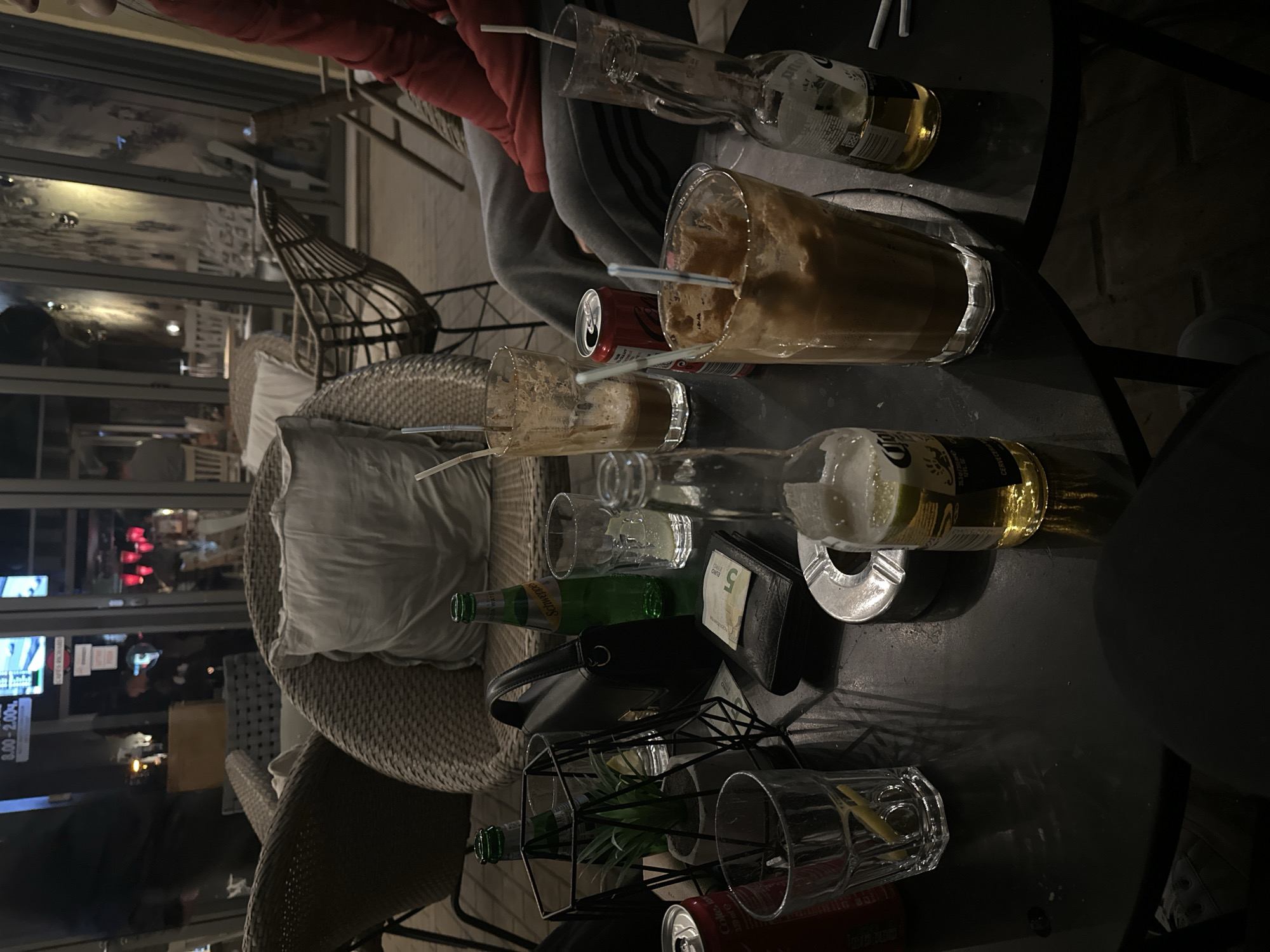The image shows a table at an outdoor seating area, possibly a cafe or restaurant. The table is littered with used glassware including empty beer bottles (Corona), a bottle of Schweppes, a few glasses with remnants of coffee or other beverages, cans of Coca-Cola, an ashtray and a black wallet containing a 5 Euro bill. A black purse is also visible on the table. The seating area has wicker chairs with cushions. In the background, there is a window reflecting an interior scene of what appears to be a storefront. Based on the store sign and exterior view reflected, the location could be in Pleven, Bulgaria. There are at least two people at the table. One is wearing a red top and the other one, a gray tracksuit.