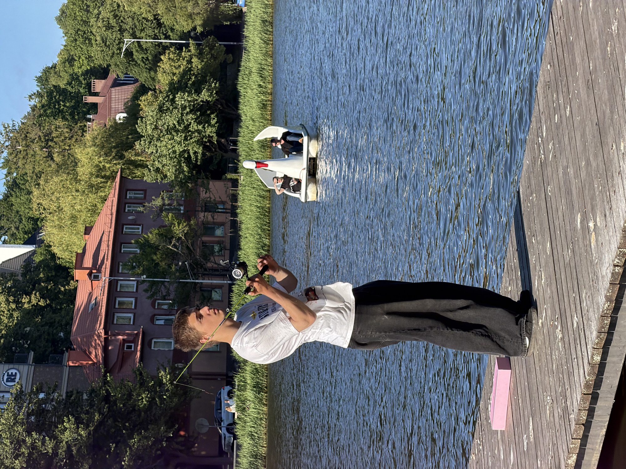 In Svetlogorsk, Russia, a young male stands on a wooden pier, fishing in a body of water. He is wearing a white t-shirt and dark pants. A pink rectangular object is on the pier near his feet. In the background, a swan-shaped pedal boat with two people is on the water. Buildings and lush green trees line the shore. The sky is clear and blue.