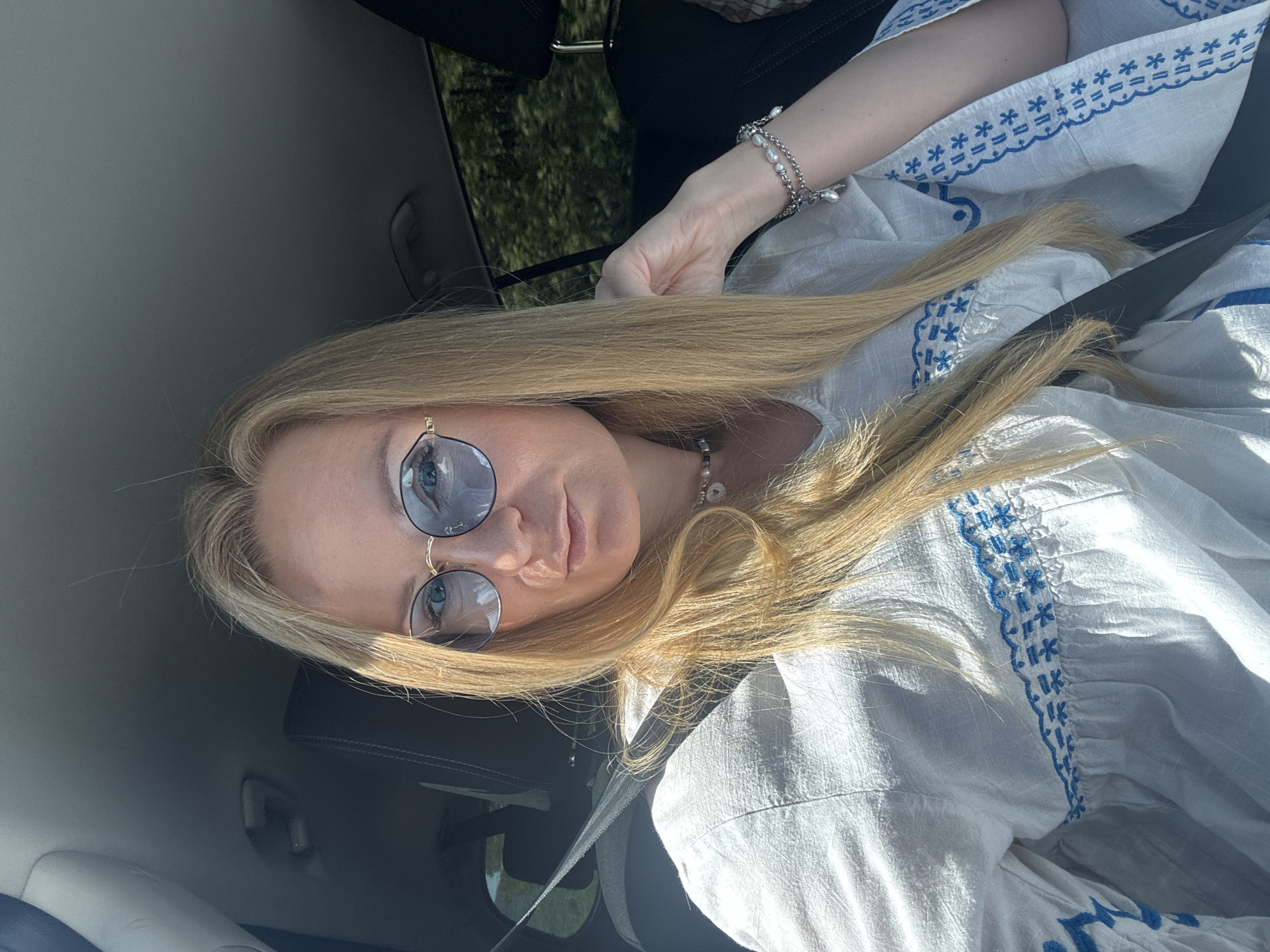 A blond-haired woman with sunglasses is in a car, wearing a white blouse with blue embroidery. She has on a bracelet and necklace with pearls. She is wearing a seatbelt. The location is Unknown, Unknown.
