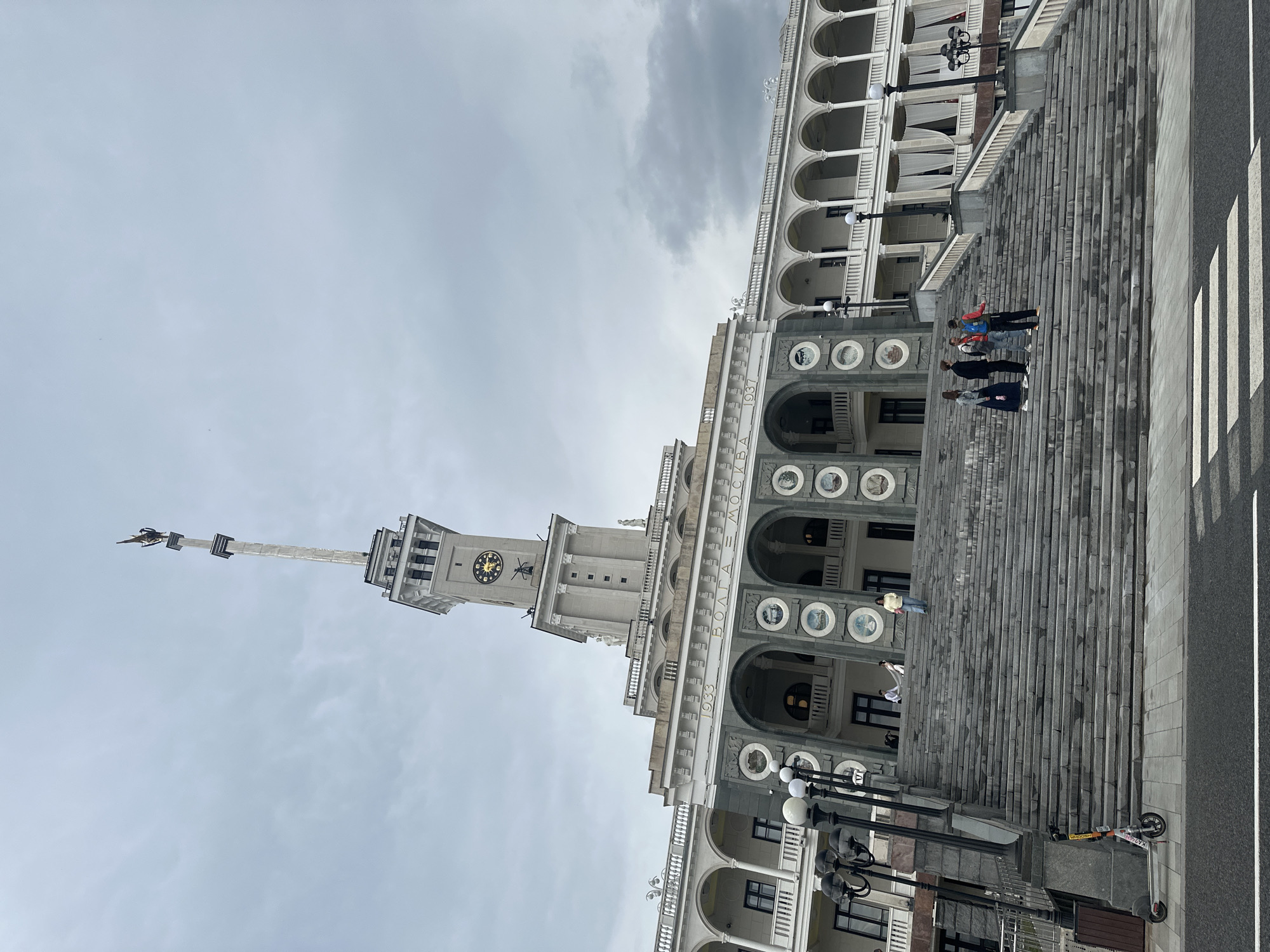The image shows a grand, ornate building with a large clock tower and a spire topped with a sculpture. The building features multiple levels of arched windows and balconies, with detailed architectural ornamentation. A wide, stone staircase leads up to the building, and several people are walking up or standing on it. The sky is overcast, suggesting a cloudy day. The overall scene conveys a sense of historical grandeur and public space.
On the building, text reads "ВОЛГА МОСКВА 1937". There are also round decorative elements with what appear to be historical scenes or portraits. On the right side of the image, a street with a pedestrian crossing is visible, and an electric scooter is parked on the sidewalk. The people in the image appear to be casually dressed tourists or visitors, with no strong emotional expressions evident. The lighting is diffused due to the cloud cover.