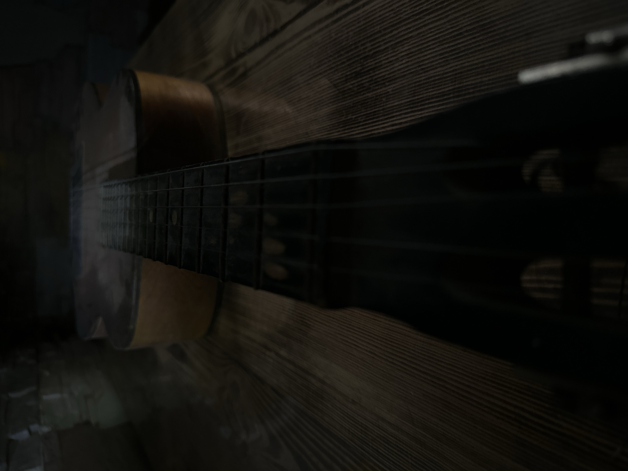 An acoustic guitar lies on a wooden surface. The guitar is positioned diagonally, with its neck extending towards the foreground and its body further away. The neck is dark and features frets. The body of the guitar is a warm, light brown wood with a glossy finish. Some soft, hazy light or smoke appears to be around the guitar. The wooden surface beneath the guitar has a visible grain pattern. The background is dark and indistinct, suggesting an indoor setting with low lighting. There are no people or other discernible objects in the image. The overall impression is one of stillness and perhaps a moody atmosphere. The location in the city of Luhansk, Ukraine, cannot be determined from the visual information provided in the image.