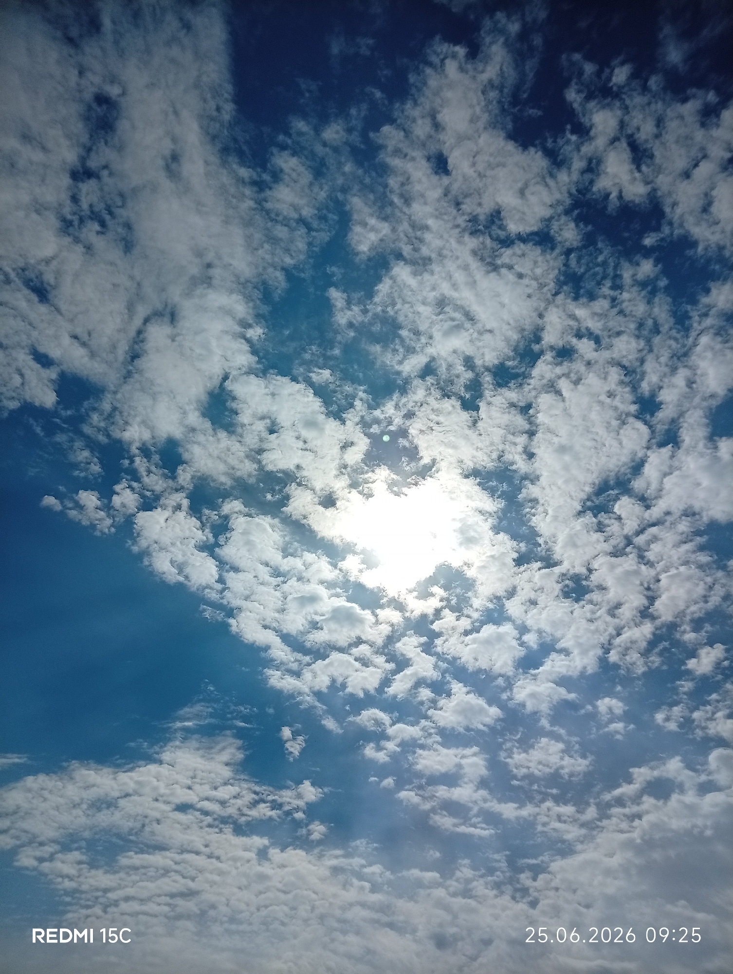 A bright, partially obscured sun shines through scattered white clouds against a clear blue sky. The image is a photograph taken outdoors, likely during the day, and features a date and time stamp in the lower right corner: "25.06.2026 09:25". The lower left corner displays "REDMI 15C", indicating the device used to take the photo. There are no people, animals, buildings, or other identifiable objects or actions visible in the frame, only the sky and clouds. The location of Mykolaivka, Ukraine, is provided as context but is not visually represented in the image itself.