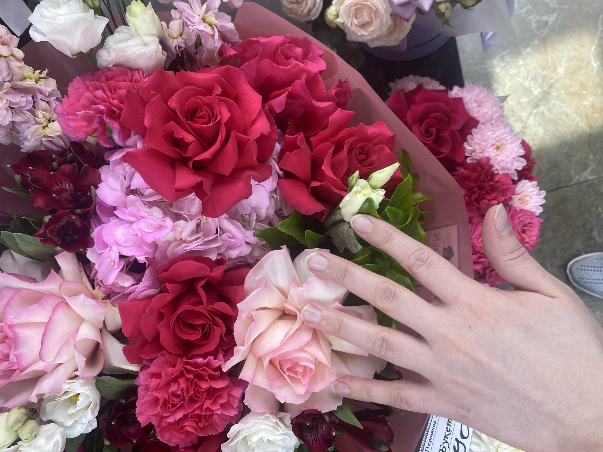 A close-up shot depicts an assortment of pink and red flowers, potentially roses and carnations, arranged in a bouquet. A Caucasian human hand is partially visible on the bouquet, fingers gently resting on the blooms. The background hints at an indoor setting. A partial Russian text can be seen printed on a tag. A section of the shoe can also be seen. Based on the printed Russian tag, the bouquet is likely to be a product of or associated with Yekaterinburg, Russia.