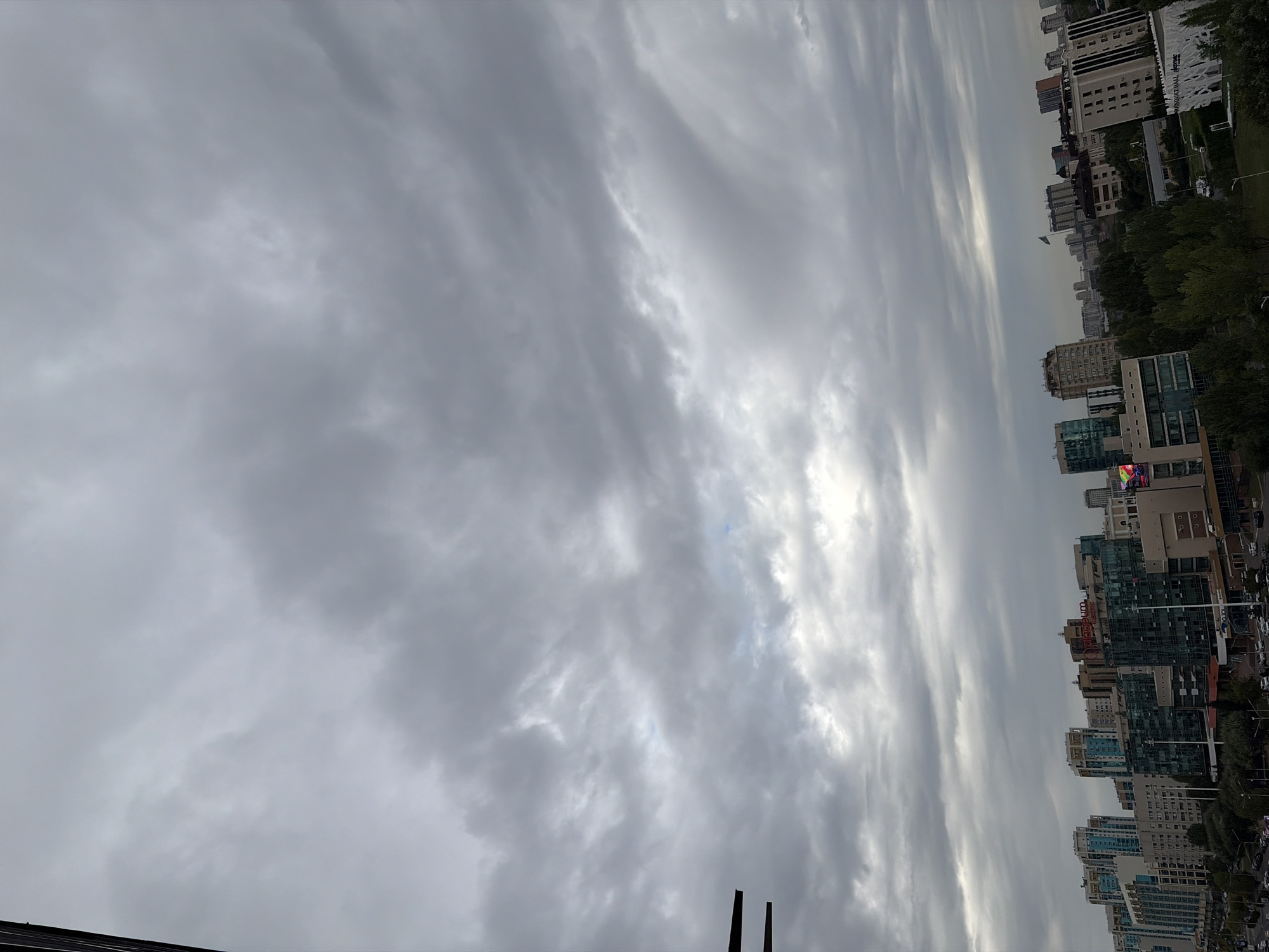 This is an outdoor, eye-level, full shot image of a city skyline under a cloudy sky, likely taken during the daytime. The city appears to be Astana, Kazakhstan, given the architecture and the flag visible in the distance.
The sky is dominated by a thick layer of grey clouds, with some lighter patches indicating diffuse sunlight filtering through. The clouds have a textured appearance, suggesting movement and variation in density.
The city below consists of a mix of modern high-rise buildings, characterized by glass facades and varied geometric shapes. Some buildings are taller and more prominent, forming the skyline, while others are shorter and appear to be commercial or residential structures. Trees line some of the streets and parks, adding a touch of greenery to the urban landscape.
Vehicles are visible on the roads, suggesting the presence of traffic, though the overall scene does not convey a sense of congestion. There are no discernible people in the image. The overall atmosphere is muted due to the cloudy weather, giving the scene a somewhat somber or calm feel.
On a tall building in the middle-left of the image, a sign with red lettering reads "Qazaqstan Electric." In the distance, a flag, likely the flag of Kazakhstan, is visible on a pole atop a building. The lighting is even, typical of an overcast day.