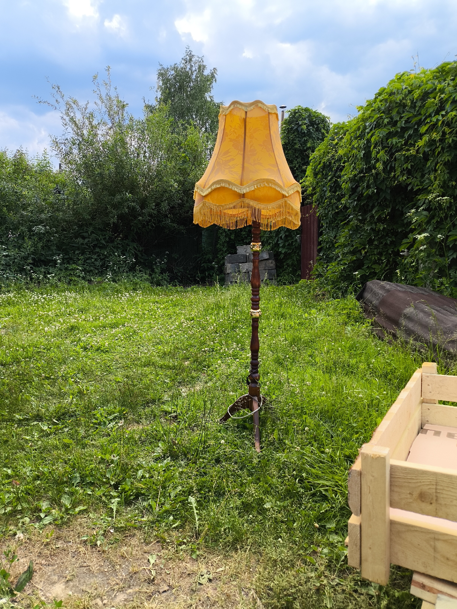 An outdoor, eye-level, wide shot captures a floor lamp standing in a grassy yard. The lamp has an ornate, orange lampshade with fringe, and its wooden base is carved with decorative elements. It is positioned in the center of the frame, with a dense green hedge forming a backdrop to the right, and a more sparse collection of bushes and trees behind the lamp to the left. The ground is covered in lush green grass, dotted with small white flowers. In the foreground on the right, part of a wooden structure, possibly a planter box, is visible. The sky above is a mix of blue with scattered white clouds.