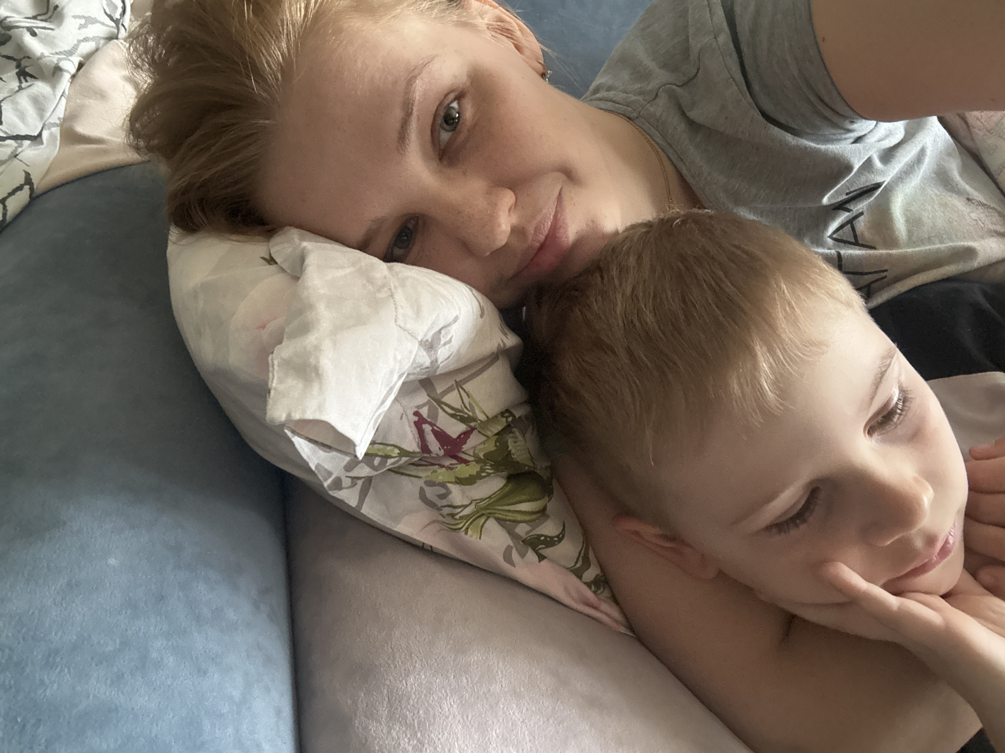 A woman and a child are lying together on a bed or couch. The woman, presumably the mother, has fair skin, reddish-blonde hair pulled back, and is wearing a gray t-shirt. She is resting her head on a patterned pillow and looking towards the camera with a slight smile. The child, who also has fair skin and blonde hair, is nestled close to the woman. The child's eyes are closed, and their hand is near their face. The background shows a blue fabric, possibly the bed or couch, and the overall impression is one of comfort and closeness. The location of the image is unknown and cannot be determined from the available information.
