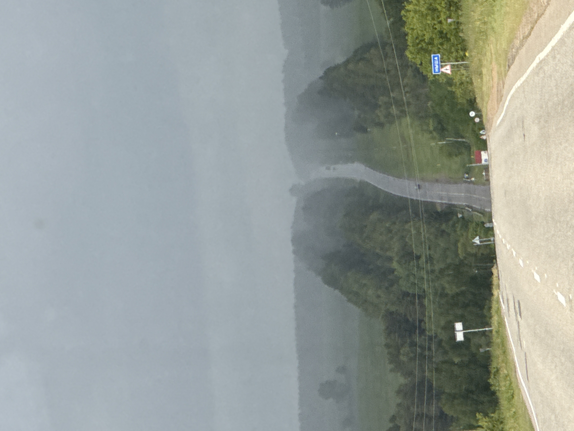 The image presents an elevated view of a rural road winding through a landscape under hazy, overcast conditions. When correctly oriented, a paved two-lane road with a solid white line on its right edge and dashed white lines in the center curves from the mid-right foreground into the distance.
The right side of the road features a grassy verge and dense green foliage, including trees and bushes, which also line the road in the middle ground. The distant landscape and the entire left half of the image are obscured by a pale, diffuse haze or overcast sky, creating a soft, muted atmosphere.
Prominently displayed on the right side of the road in the mid-ground is a blue rectangular road sign with white text clearly reading "Ныша" (Nysha). Next to it is a smaller white triangular warning sign with a red border. Power lines run parallel to the road further into the scene. Other small, indistinct road signs and structures are visible along the roadside in the distance. The scene suggests a quiet, potentially foggy or damp day in a remote area, with no people or active vehicles clearly visible.