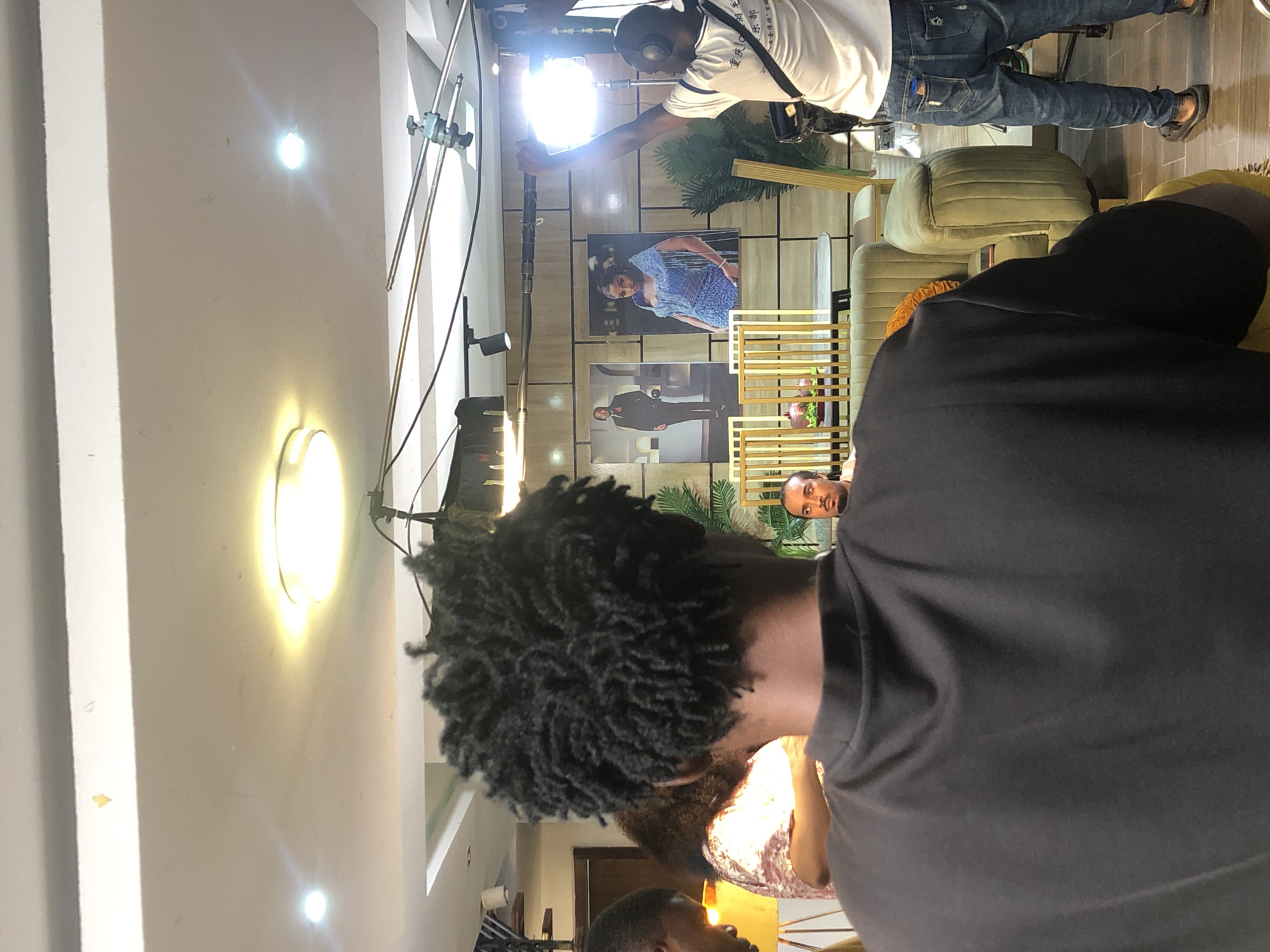 The image depicts an indoor scene in Unknown, Unknown, possibly a studio or event space, focused on a behind-the-scenes moment during what appears to be a photoshoot or filming session. In the foreground, the back of a person with short, dark, tightly curled hair is visible, wearing a dark gray shirt. Behind this person, a man with facial hair is seated and looking at the camera, with a glimpse of an orange patterned garment around his shoulders. In the middle ground, two large framed photos are mounted on the wall, one of a woman in a blue dress and another of a man in a suit. A person in a white shirt and jeans is holding a camera and adjusting a bright light source on a stand. The background shows modern interior design elements, including gold decorative structures, potted plants, and upholstered furniture. Bright ceiling lights illuminate the space.
