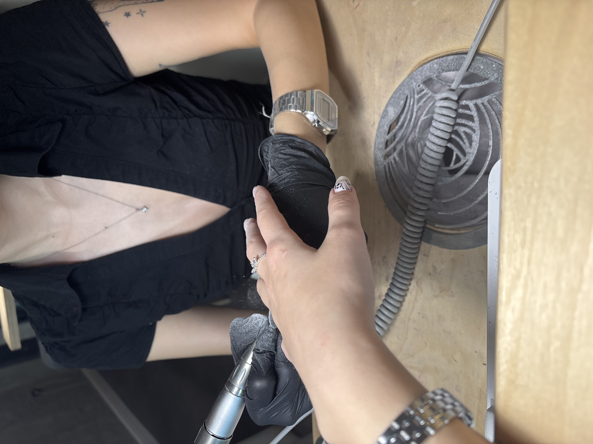 A person wearing a black shirt, black gloves, and two different wristwatches is working on nails with a professional electric nail drill. The person is likely a nail technician providing a service, as they are wearing protective gloves and using specialized equipment. The environment appears to be a nail salon or a similar setting, with a gray ventilation system on the table, typically used to extract dust and fumes during nail procedures. There is no information in the image to suggest it is located in Sofia, Bulgaria.