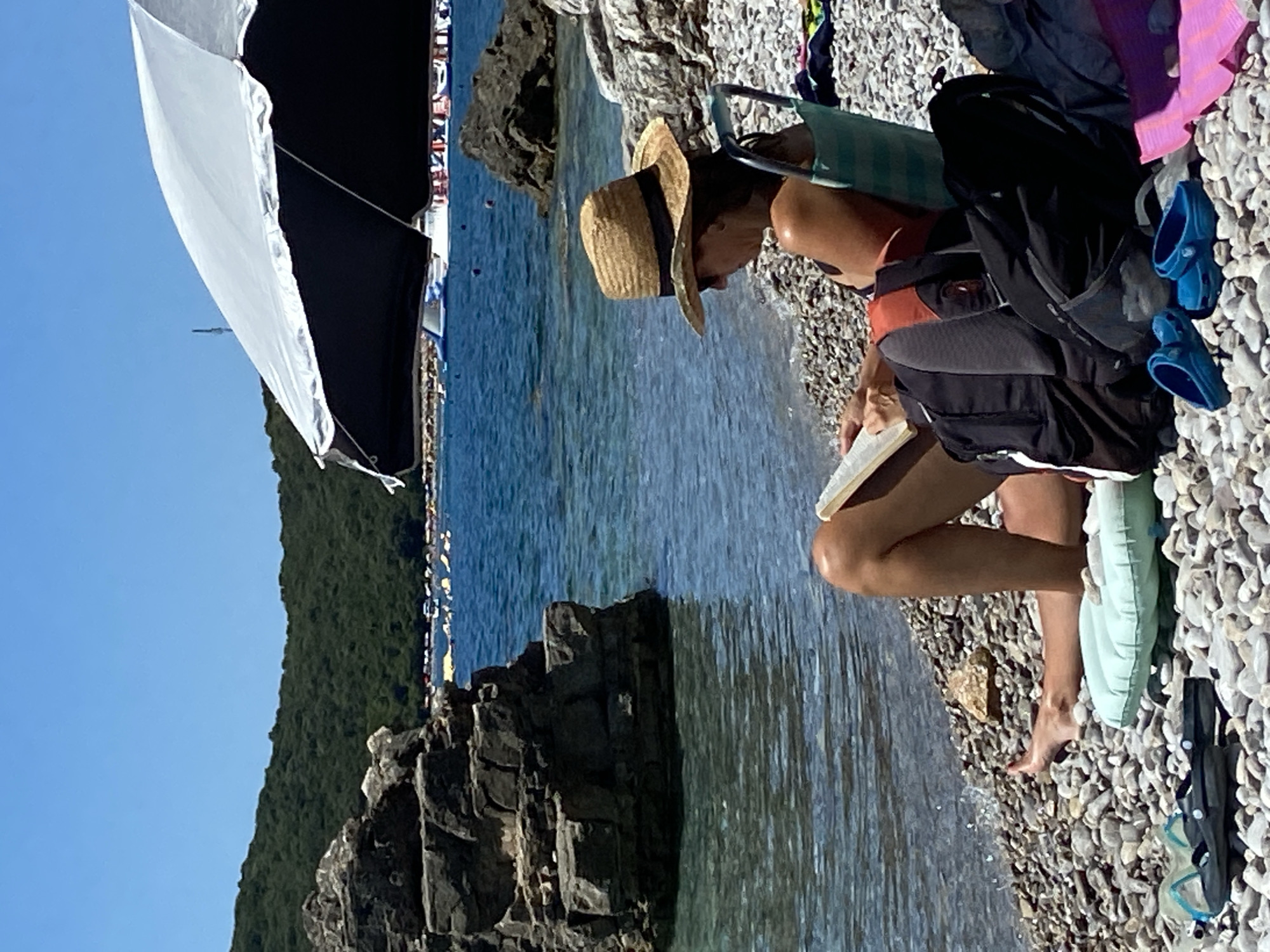 In the city of Prijevor, Montenegro, on a sunny day, a woman sits on a pebble beach, reading a book. She is wearing a straw hat and a swimsuit, and is seated in a folding chair. Next to her are two backpacks, a pair of blue Crocs, and an inflatable pillow. The beach is next to the clear blue water of the sea, with a rocky hill and green vegetation in the background. There are several inflatable water attractions and boats visible on the water in the distance, suggesting a popular tourist area. A large umbrella provides shade for the woman.