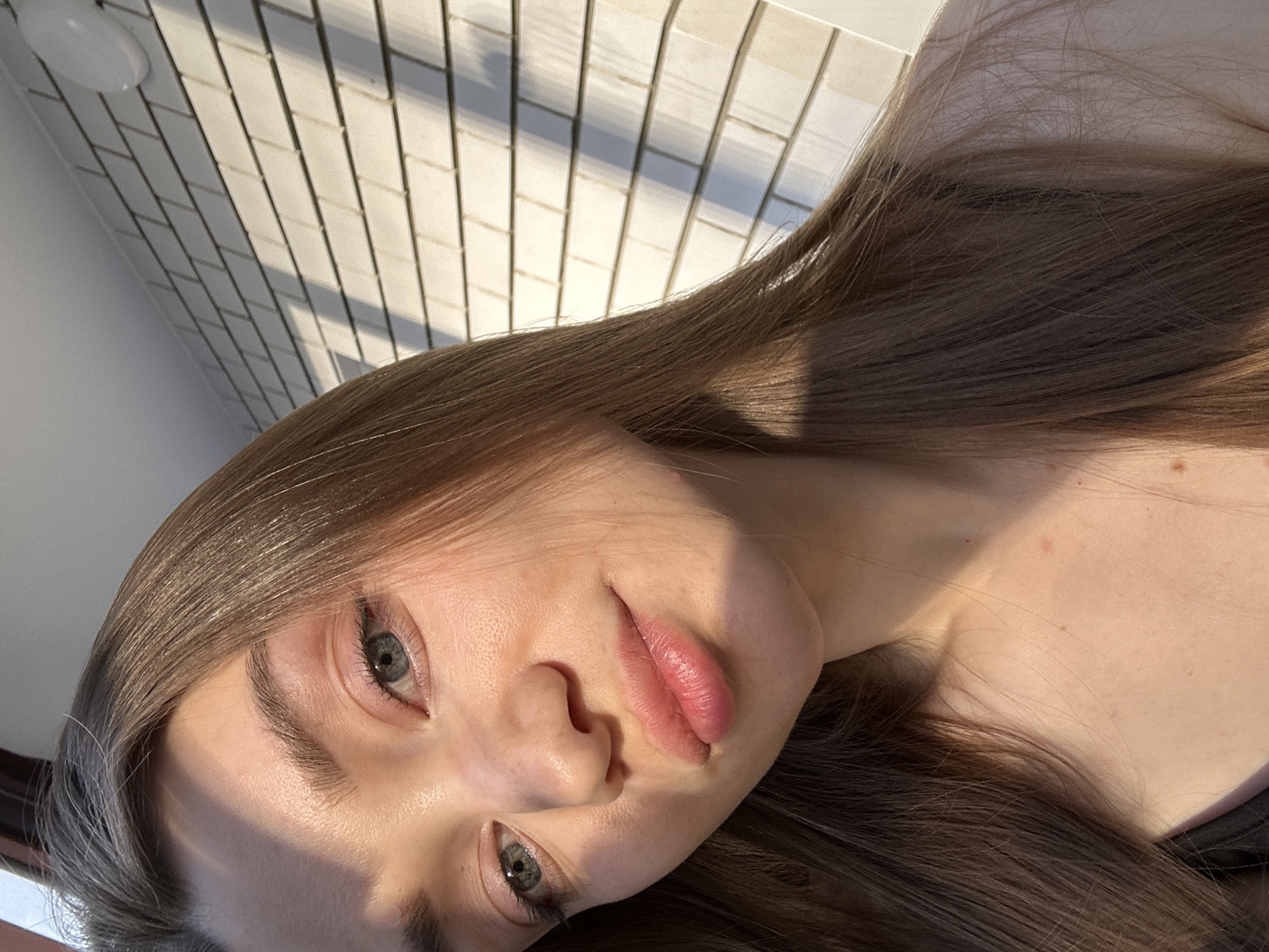 A close-up, tilted portrait features a young woman with long, straight brown hair. Her face, illuminated by strong, direct sunlight, shows light blue-grey eyes, a nose, and full, light pink lips. Distinct shadows fall across her forehead, under her eye, and along her cheek and nose, indicating the angle of the sun. She looks directly towards the viewer with a neutral expression.
The background consists of a white tiled wall with a staggered rectangular pattern, also showing prominent, elongated shadows that match the lighting on the subject. A small, round white light fixture is visible in the upper left corner of the background. A dark strap of clothing is partially visible on her lower right. The scene is lit by natural light, suggesting either morning or late afternoon sun. This casual moment is captured indoors, potentially in Kazan, Russia. No text is visible.