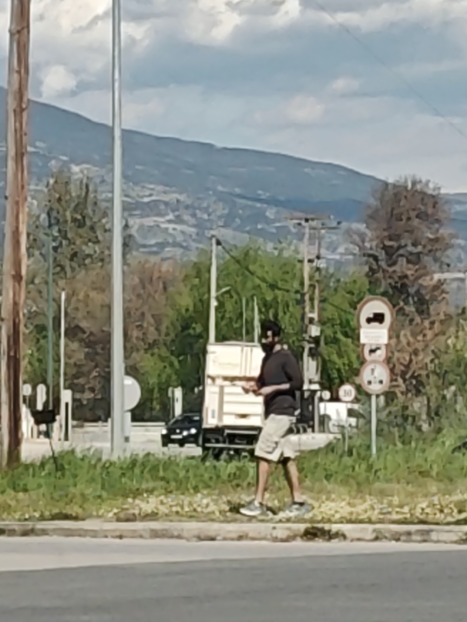 The image shows a man walking on a sidewalk next to a road in what appears to be an urban area, potentially the city of Serres, Greece, given the context. He is wearing a dark shirt, light-colored shorts, and sneakers, and he is wearing a face mask. He is holding an object in his hands, although the specific nature of the object is unclear.
In the background, there is a large truck parked next to the sidewalk with its back door open. Behind the truck are green trees and hills. In the distance, a mountain range is visible under a cloudy sky. Road signs are positioned near the truck. There is a black car that is approaching the truck.