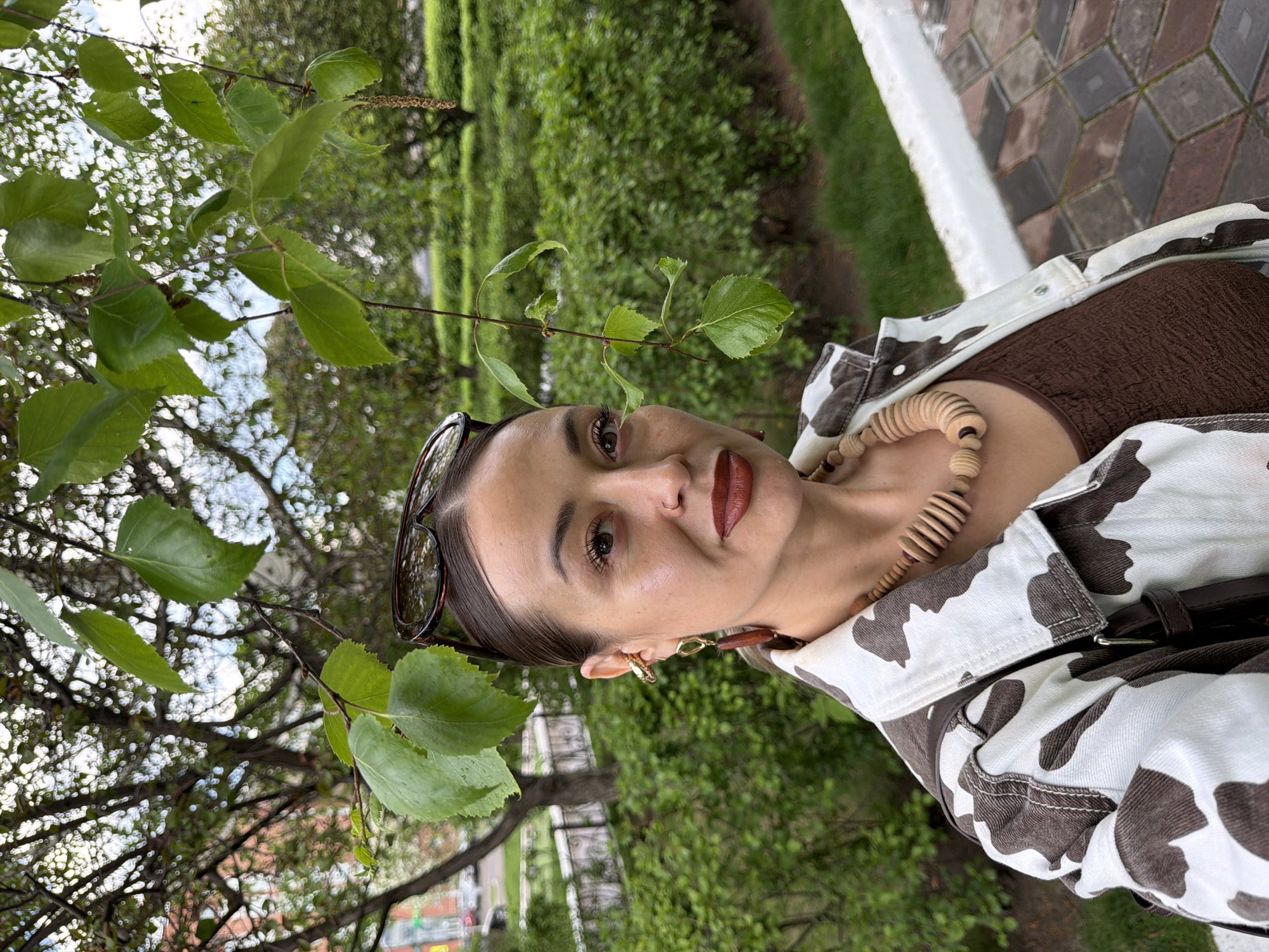 A woman is standing outdoors, likely in a park or garden setting based on the lush green foliage behind her and to her left. She is wearing a brown top, a white and brown cow-patterned jacket, and a wooden bead necklace. Sunglasses are perched on her head. Her makeup includes brown lipstick. She is near tree branches with leaves, some of which are wet. The backdrop includes trimmed hedges and trees. The image appears to be taken near an urban environment. It is not possible to confirm the exact location of Krasnoyarsk, Russia, based solely on this image.