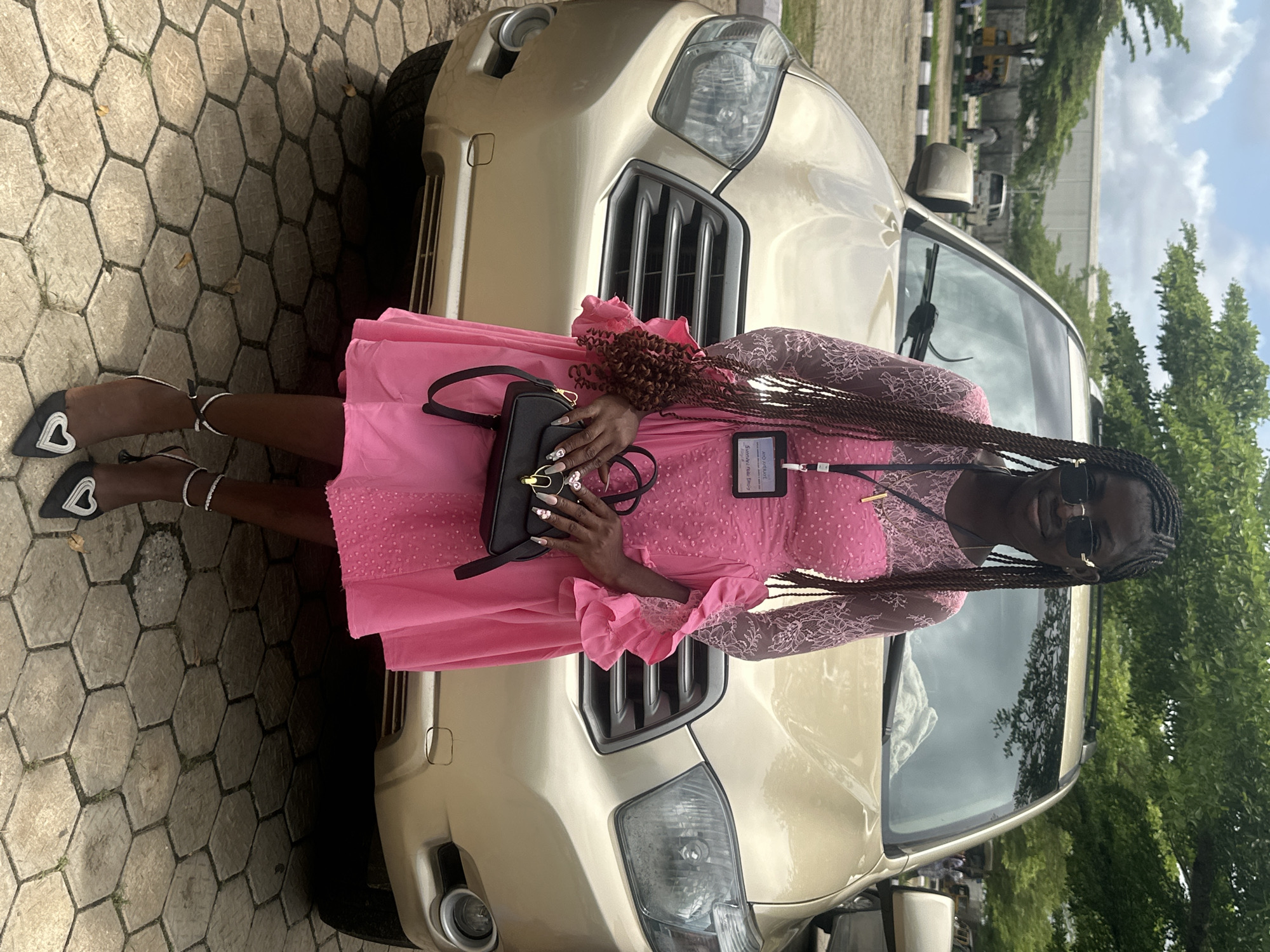 A young Black woman stands in front of a gold SUV. She is wearing a pink dress with lace sleeves and a ruffled hem, black high heels with heart-shaped embellishments, and a black handbag. She has long braided hair and is wearing sunglasses. The vehicle is parked on a paved area in what appears to be a public space, possibly an event venue or parking lot in Itoga, Nigeria. Trees and other vehicles are visible in the background, suggesting an outdoor setting within the city. The overall impression is of a formal occasion or outing.