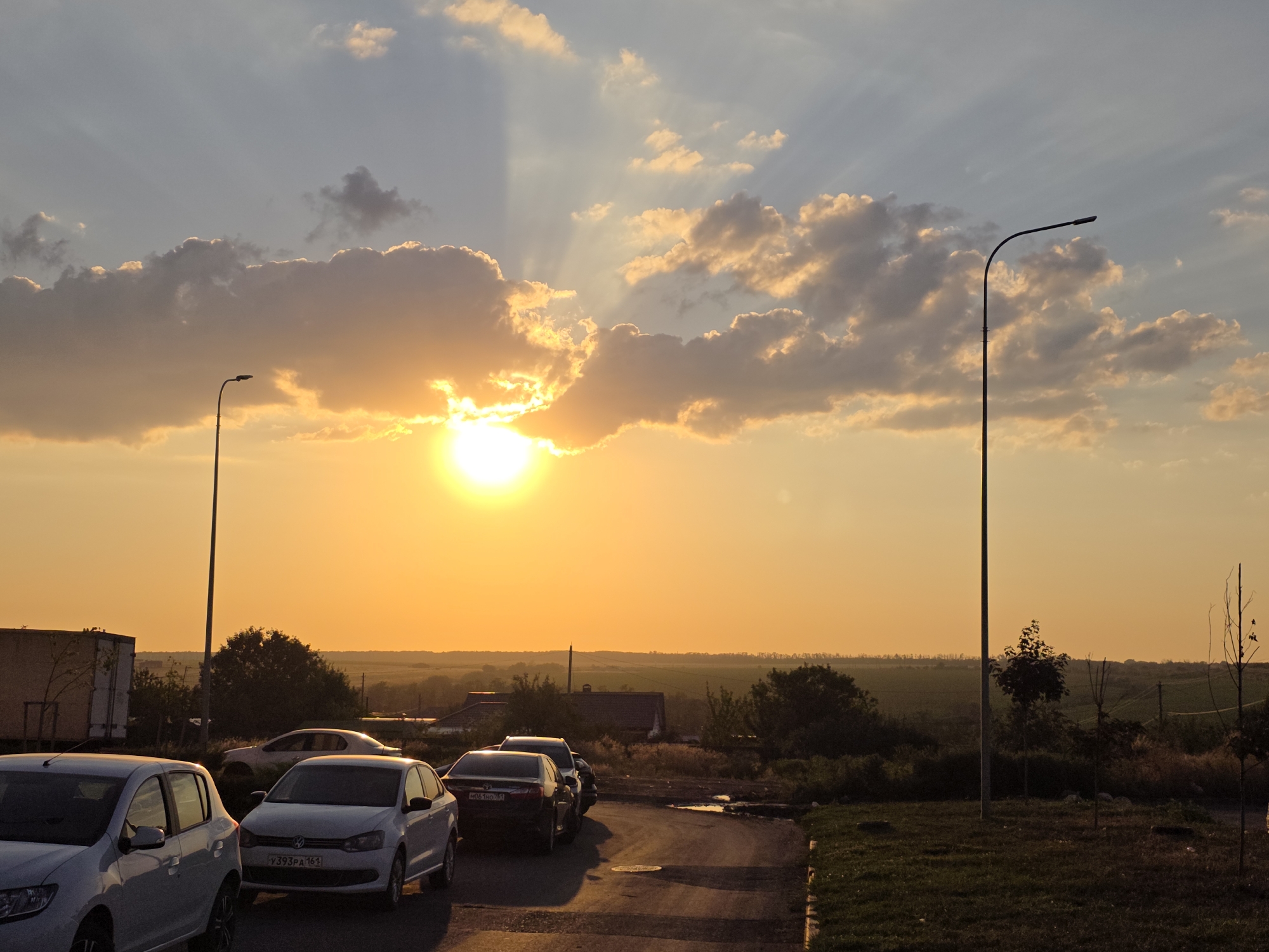 The image captures a sunset over Rostov-on-Don, Russia. The sun is low in the sky, partially obscured by clouds, casting a warm orange glow. Several cars are parked on a paved surface in the foreground, with a white truck visible to the left. In the background, there is a landscape of fields and trees, with houses scattered throughout. Two streetlights are present in the scene, one on the left and another on the right. The sky is a mix of blue and orange, with dramatic cloud formations. The overall impression is one of a peaceful rural evening.