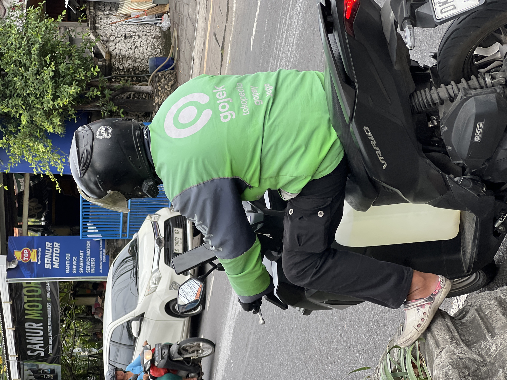 In Denpasar, Indonesia, a Gojek driver, identifiable by their bright green jacket with the Gojek logo, is seen from behind on a black motorcycle. The driver wears a black helmet, black pants, and a pair of light-colored Crocs with pink and white accents. They are seated on the motorcycle, which has a clear plastic container strapped to the rear. The motorcycle's license plate is partially visible. In the background, a white car is parked near a business with signage that includes "SANUR MOTOR" and "TOP Synthetic Oil". A woman on another motorcycle is also visible in the left background. The scene appears to be on a street with paved asphalt.